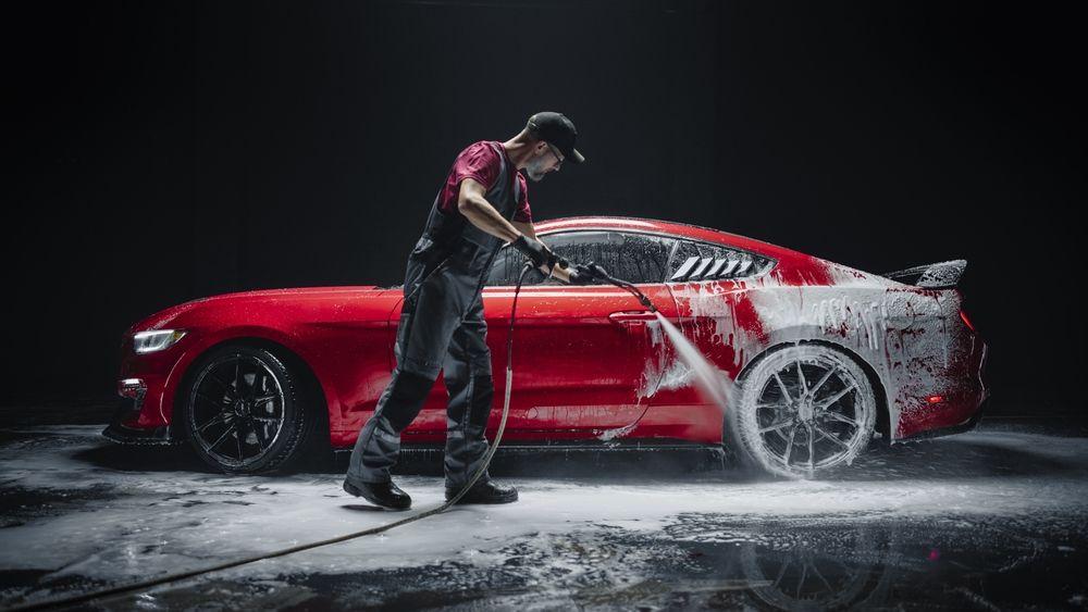 Man washing a red sports car with a pressure washer, black background.