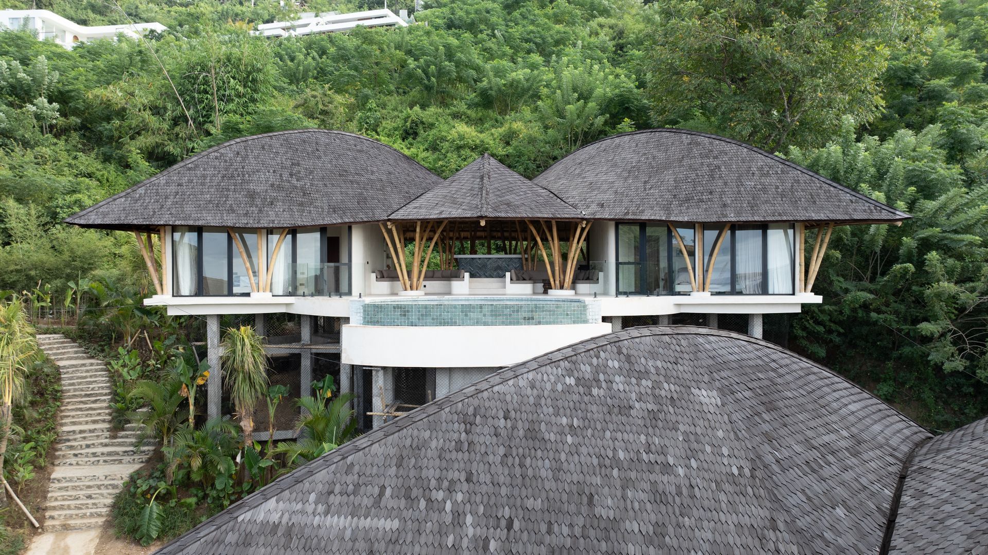 Luxury villa with thatched roofs, bamboo supports, and a pool, nestled in a lush green hillside.