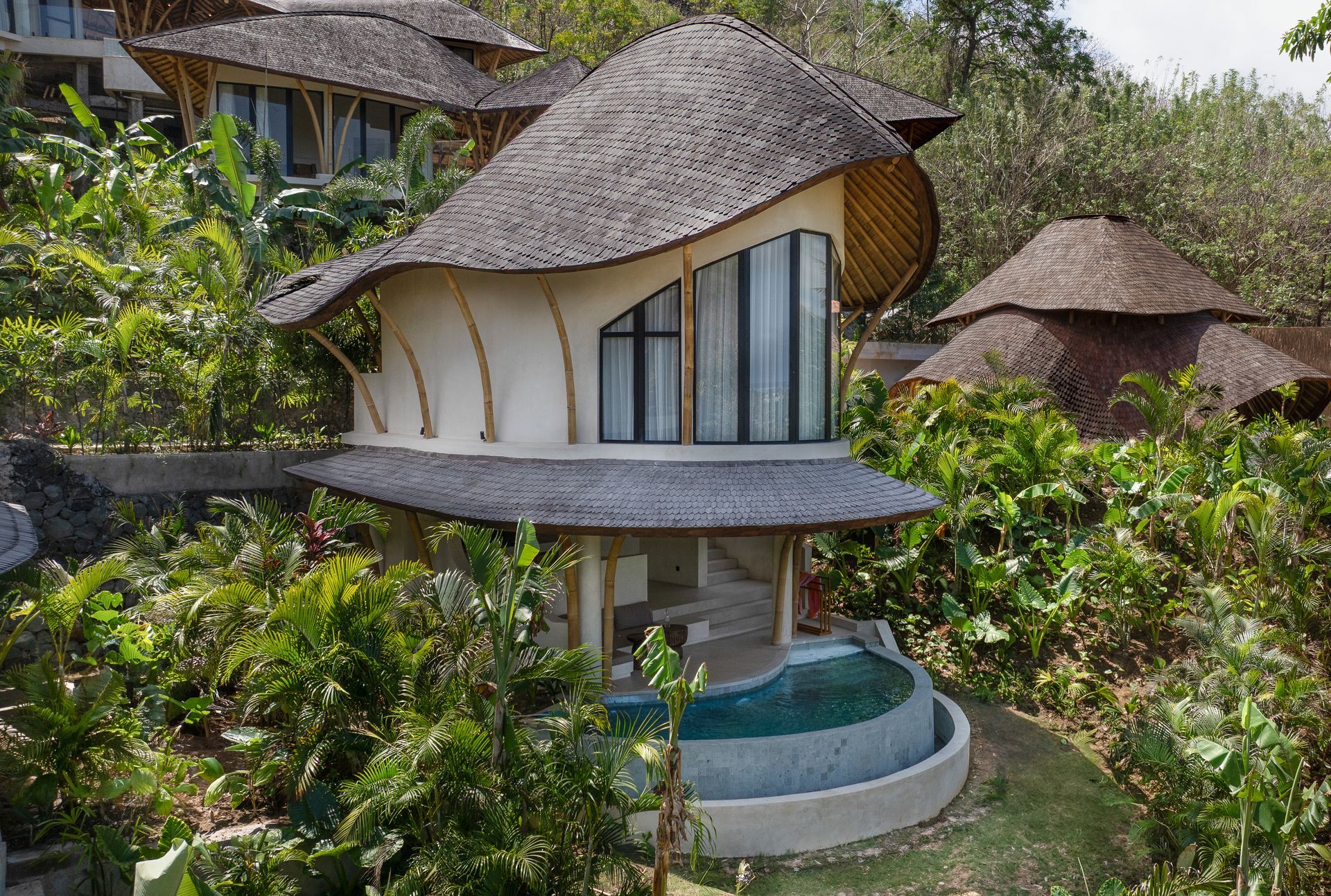 Whimsical house with a thatched roof, surrounded by lush greenery and a small pool.