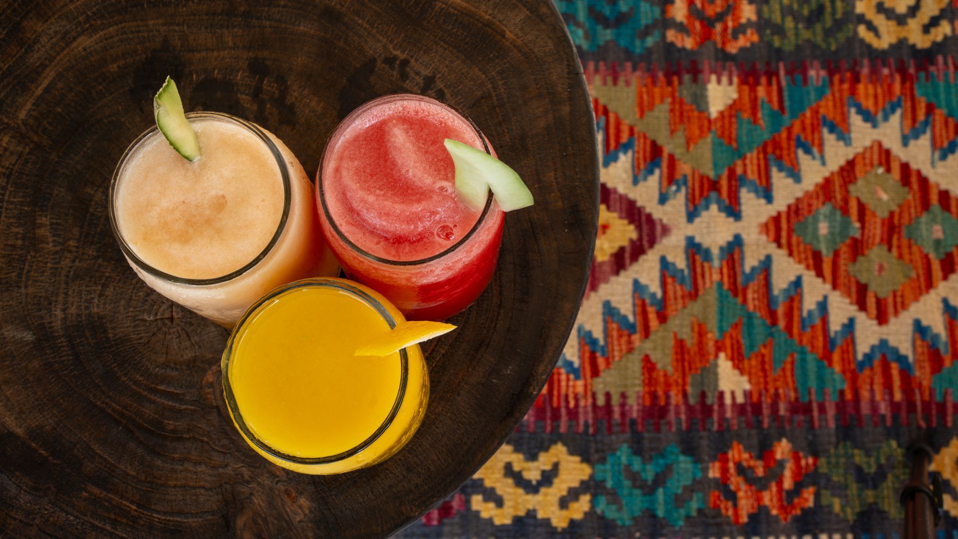 Three colorful fruit smoothies in glasses, garnished with fruit slices, on a wooden surface, with a patterned rug.