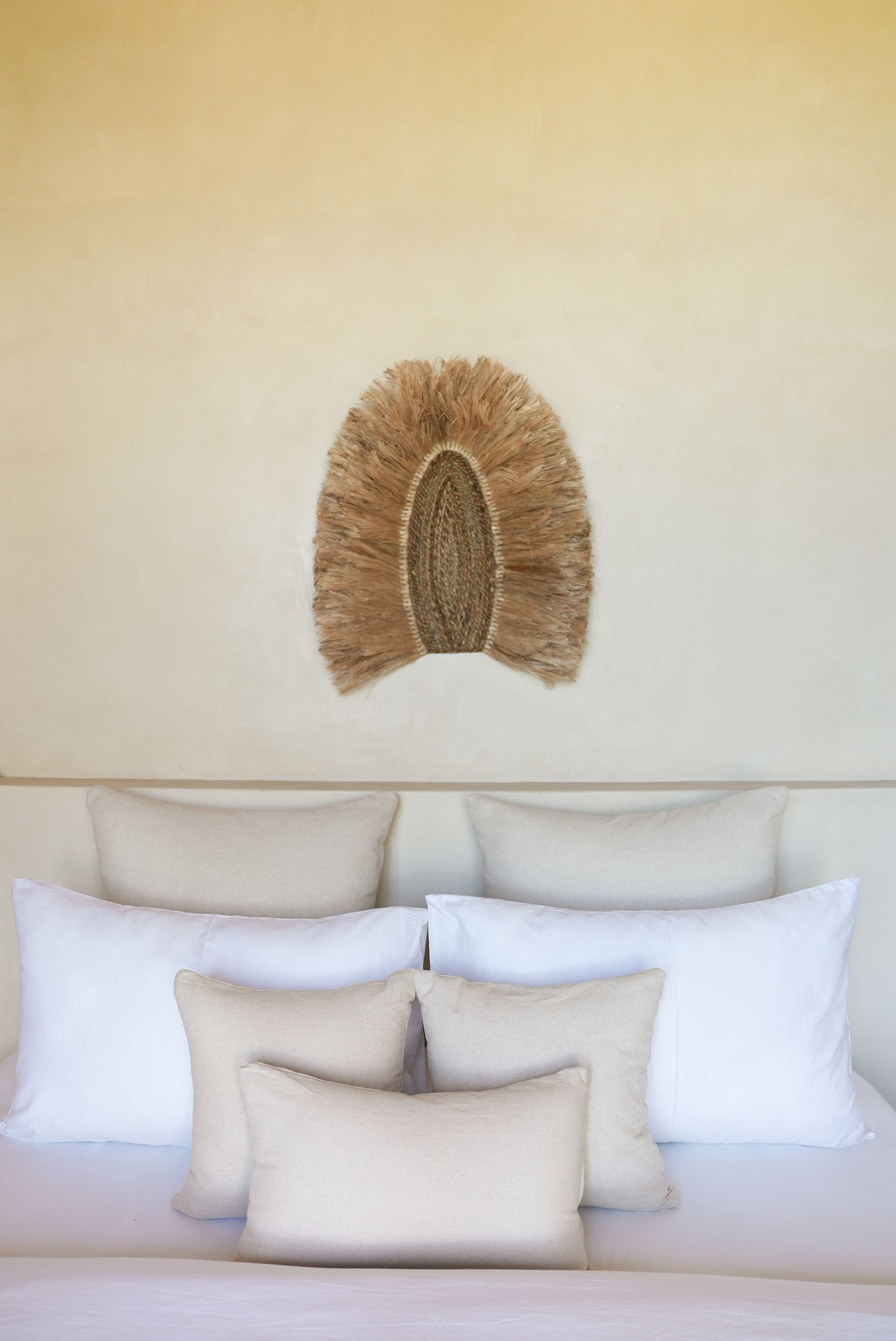 Bed with beige and white pillows, decorated with a woven wall hanging.