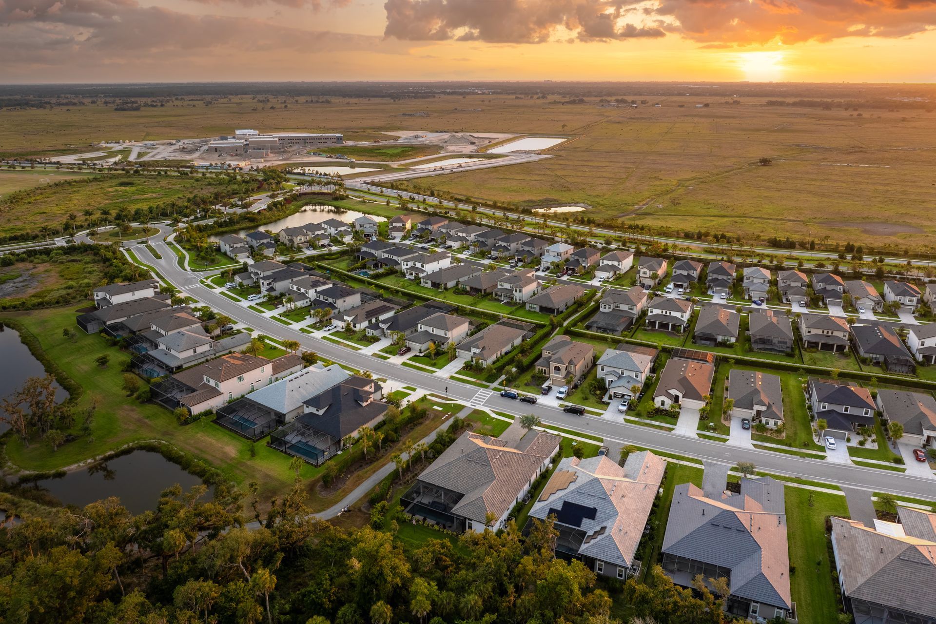 Aerial view of a suburban neighborhood at sunset, with rows of houses, roads, and a large natural area in the background.