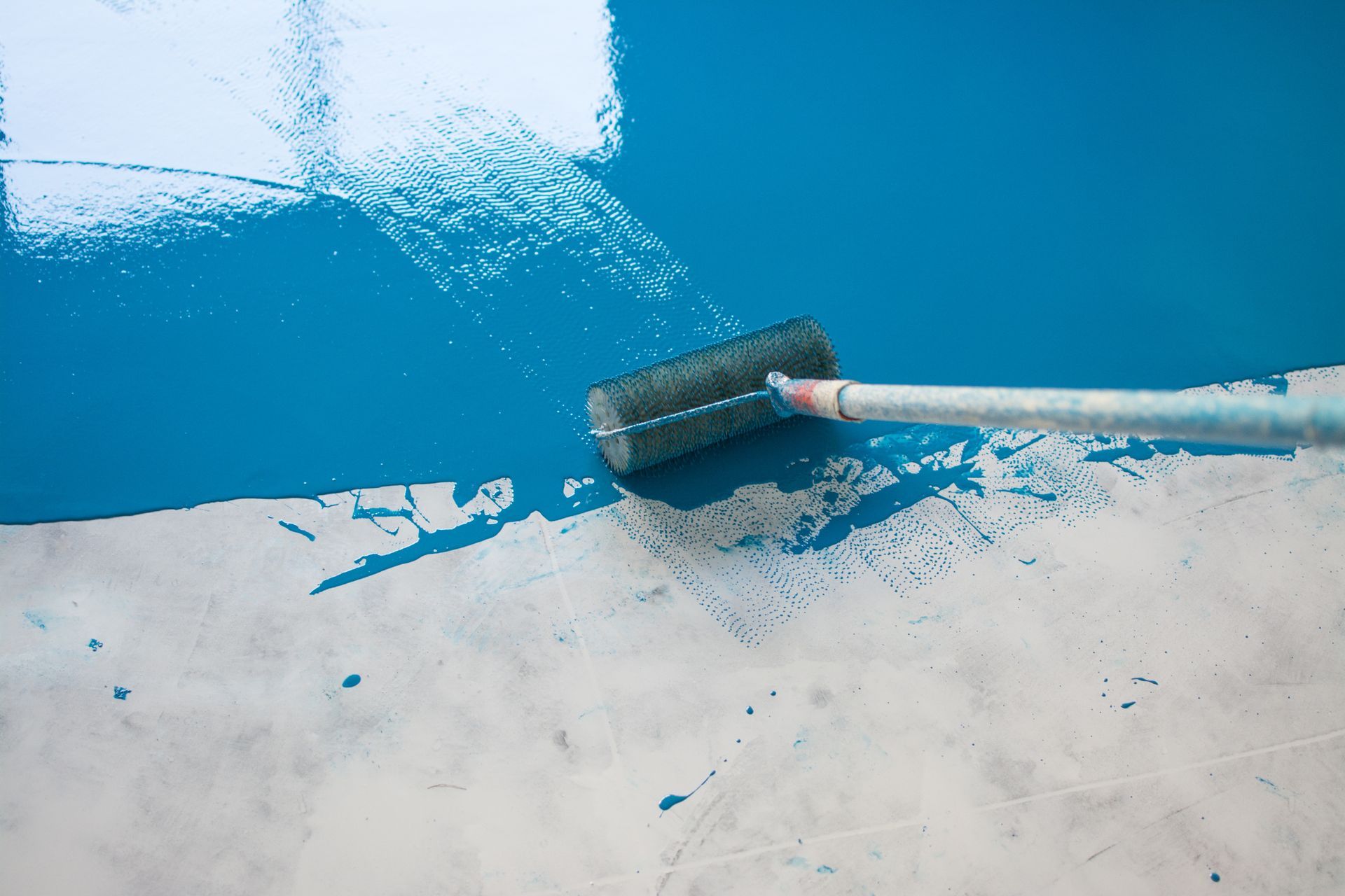 A paint roller applying blue paint to a concrete surface, with areas already painted and unpainted.