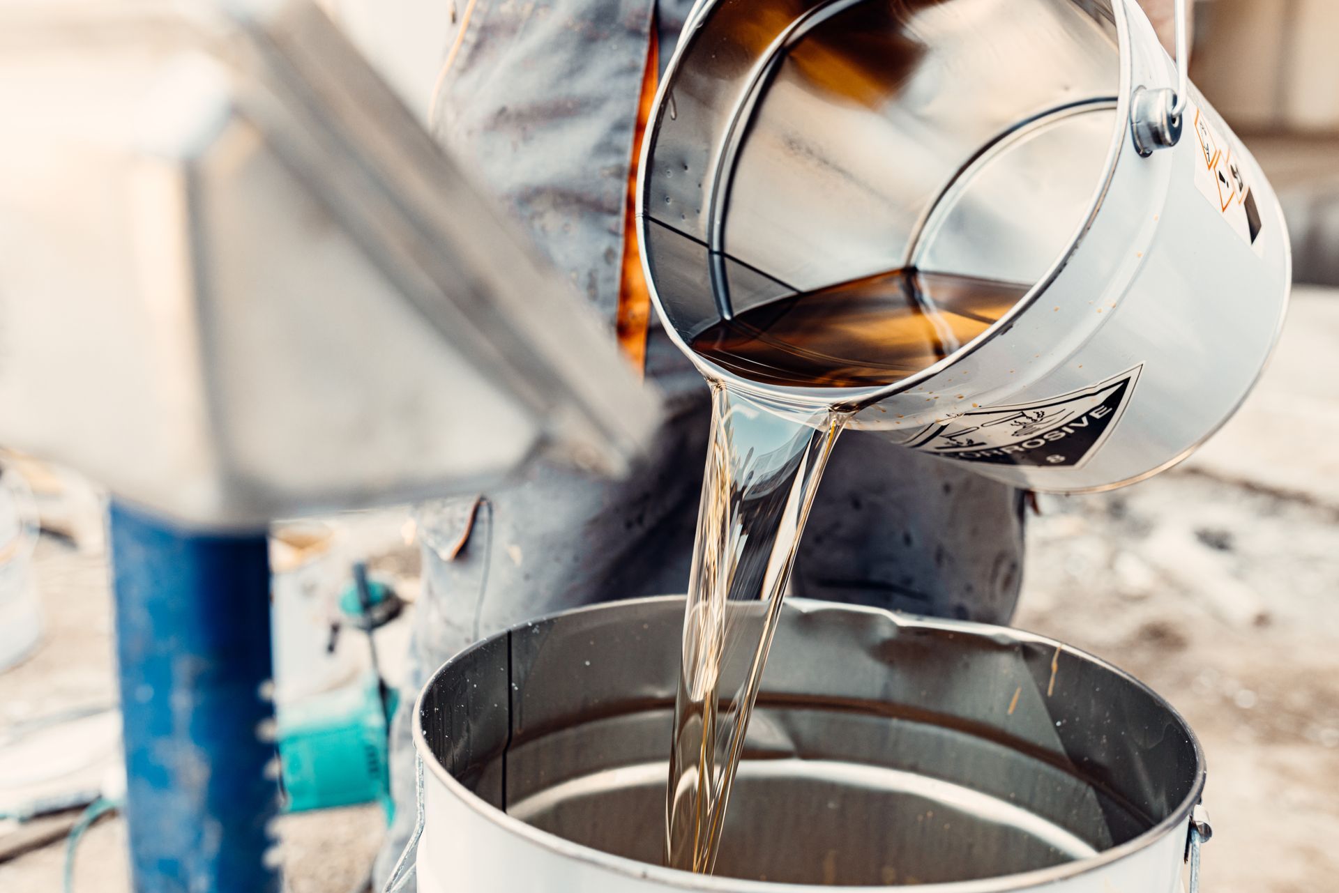 Worker pouring liquid from a metal bucket into a larger metal container.