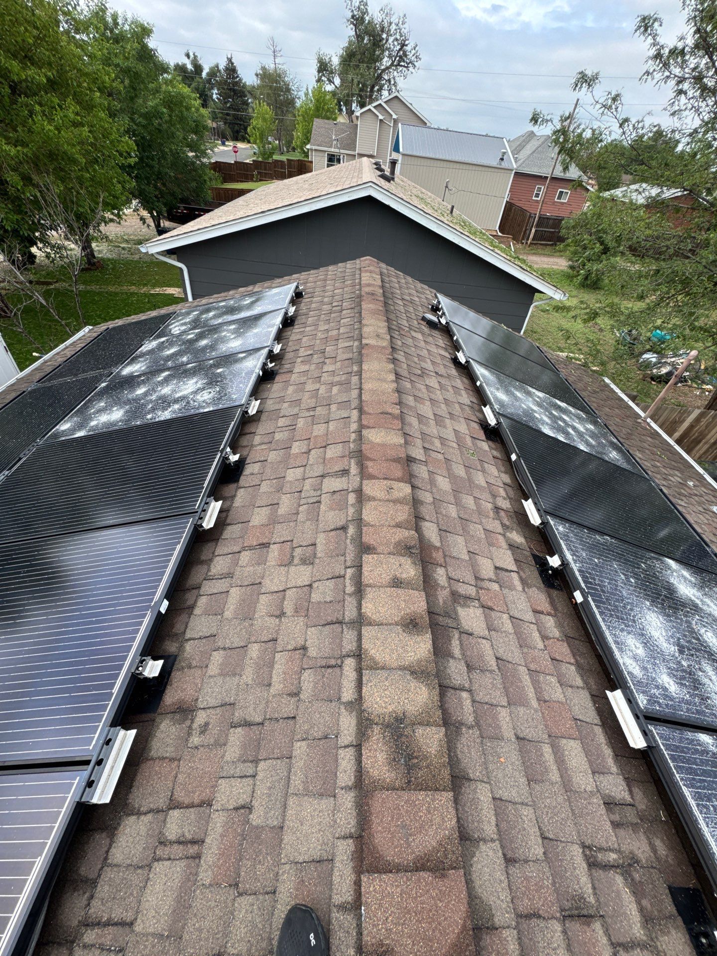 Solar panels installed on a brown shingled roof, looking up towards a house against a cloudy sky.