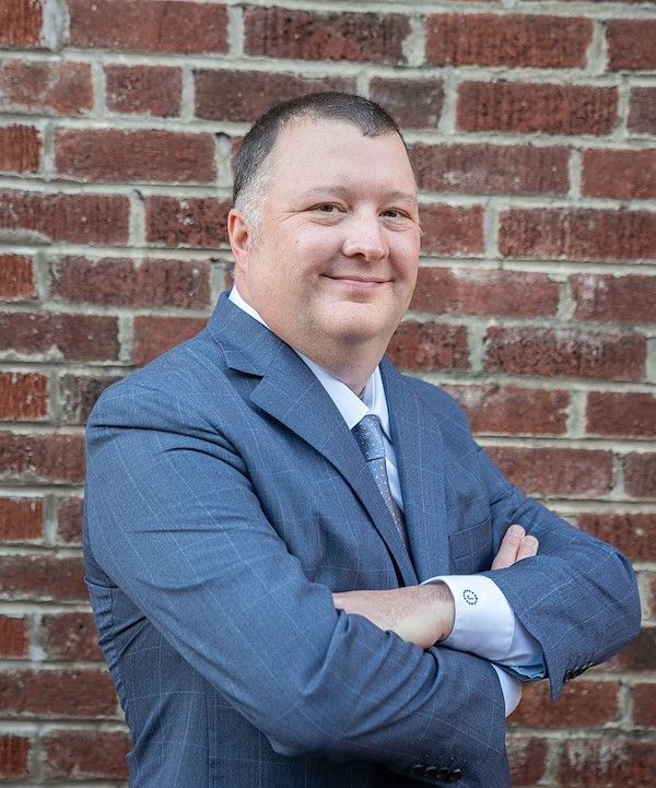 Man in a suit smiles, outdoors. Light blue tie, checkered shirt, green bushes and pink flowers in the background.