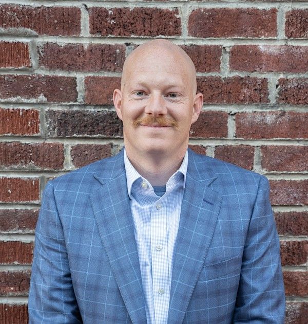 Man with shaved head leans against a brick wall, arms crossed, wearing a tie and collared shirt.