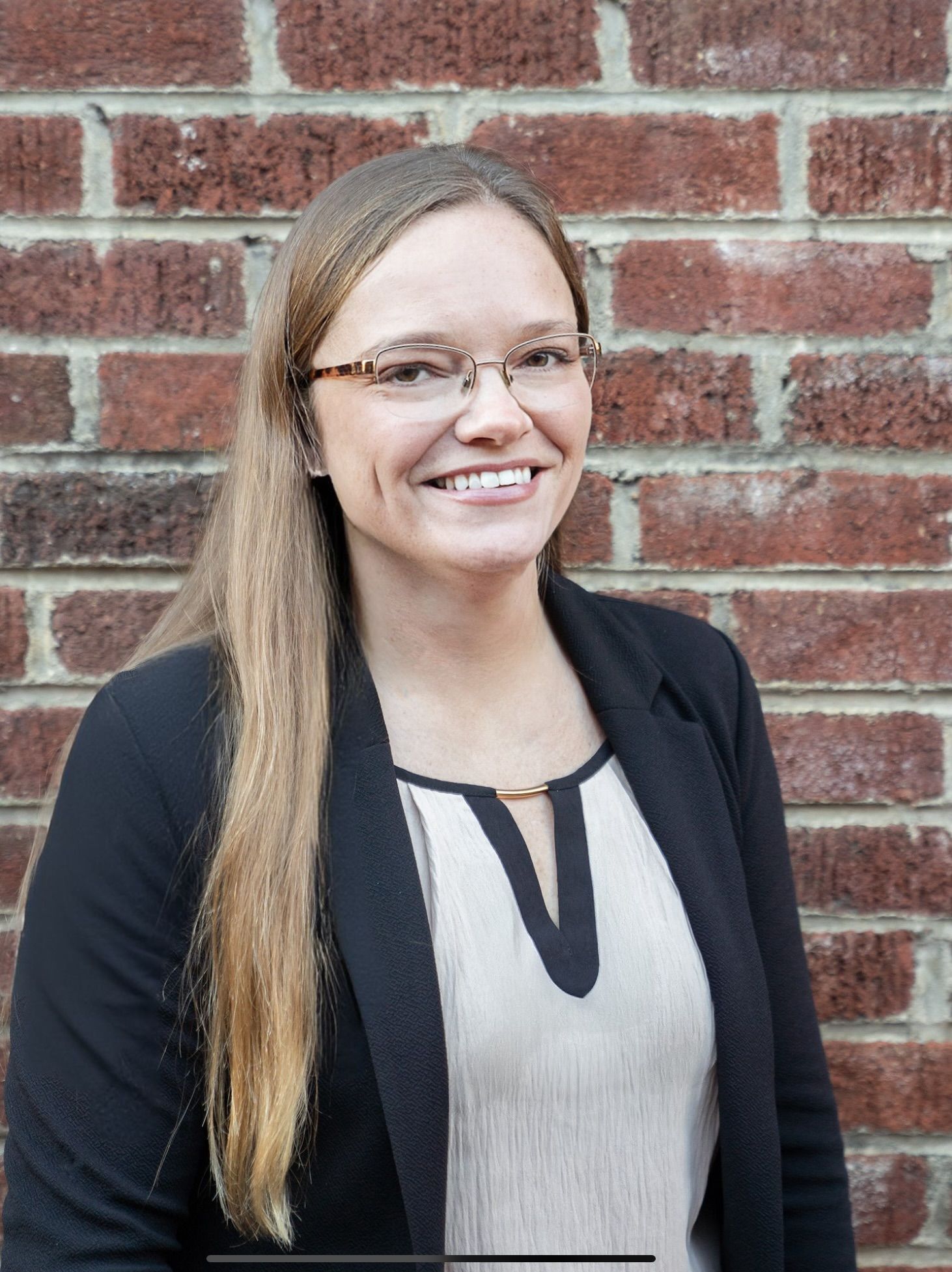 Woman with glasses smiles while sitting in a black chair. She wears a floral print top with a cross neckline.