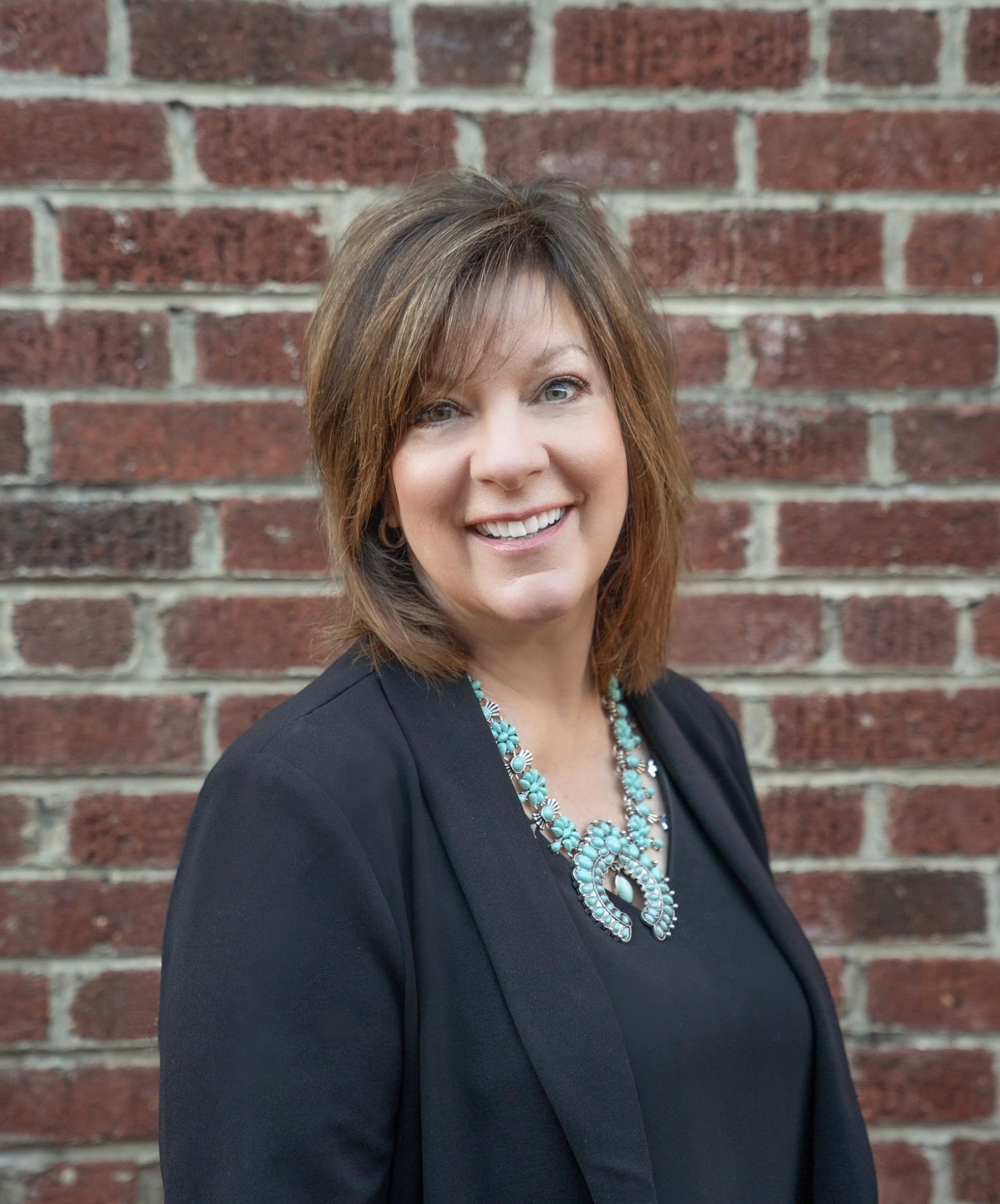 Woman in a light blue blazer, leaning on a black leather chair, smiling.