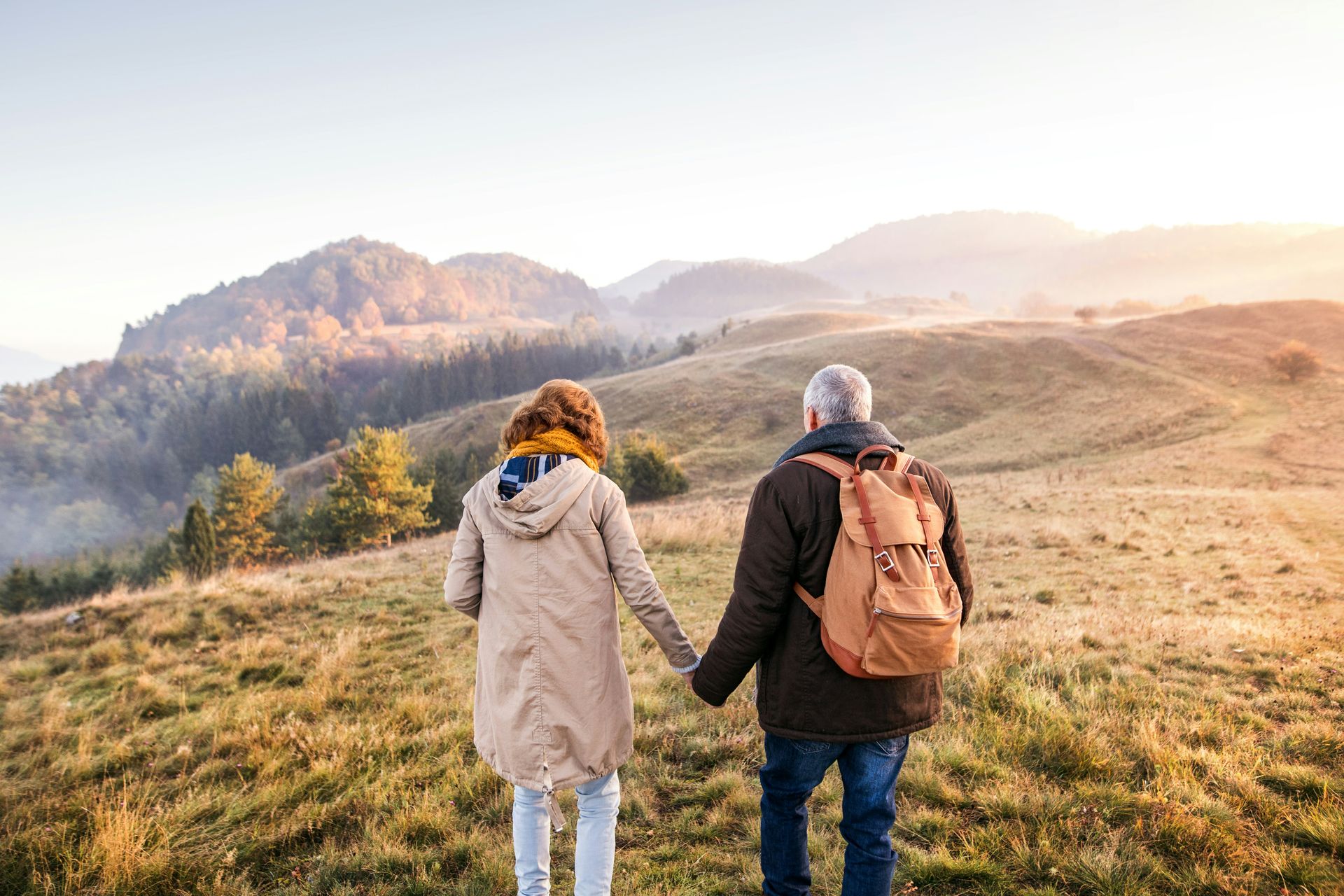Couple holding hands, walking on a hilltop with a backpack, overlooking mountains in golden sunlight.