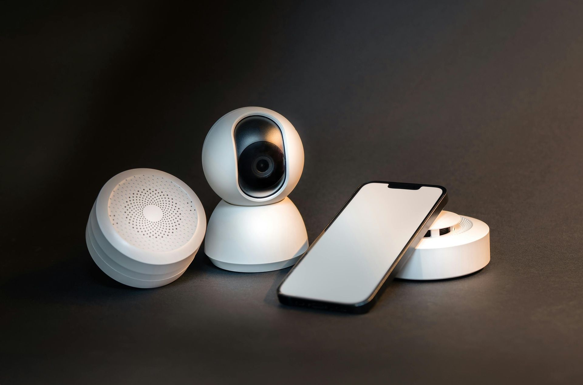 White security camera, smart speaker, and smartphone arranged on a dark surface.