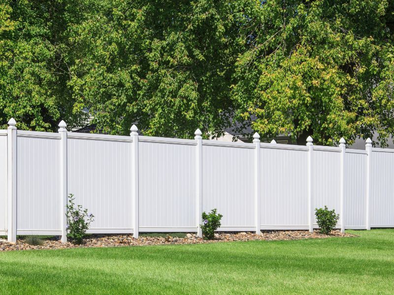 White vinyl fence in a grassy yard, with green trees in the background.