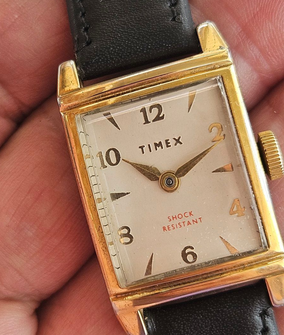 Gold rectangular Timex watch with black leather strap on a person's hand. Beige face with black numerals, red 
