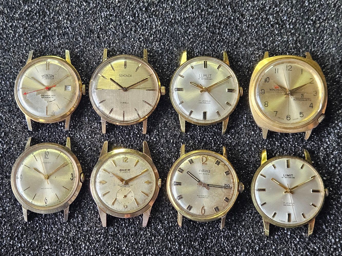 Eight vintage gold-tone wrist watch faces arranged on a textured dark surface.