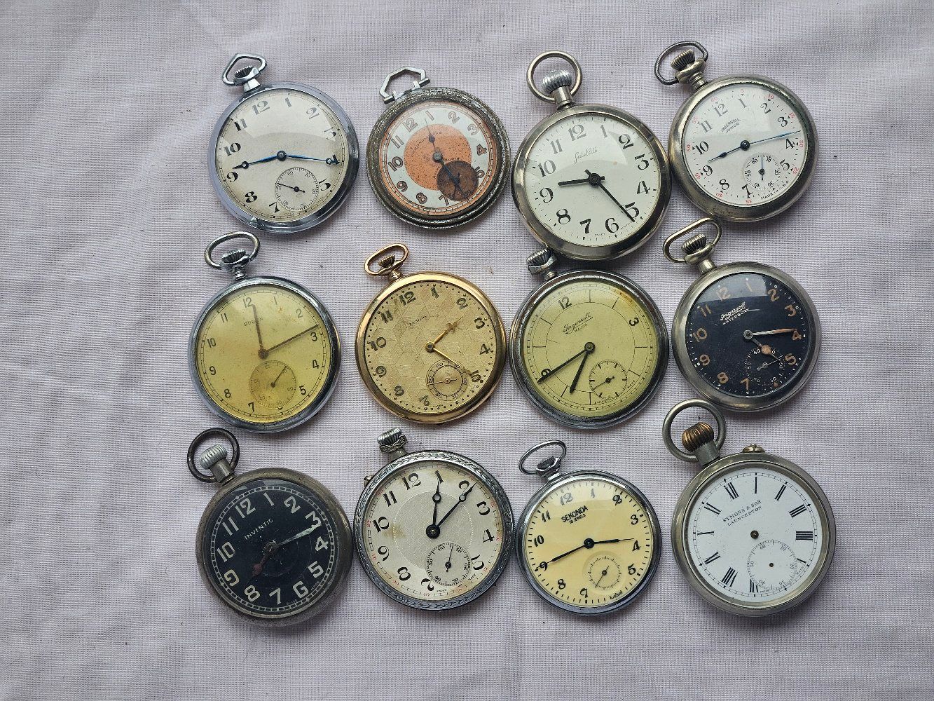 Twelve antique pocket watches on a textured surface, each with a different face and metal casing.