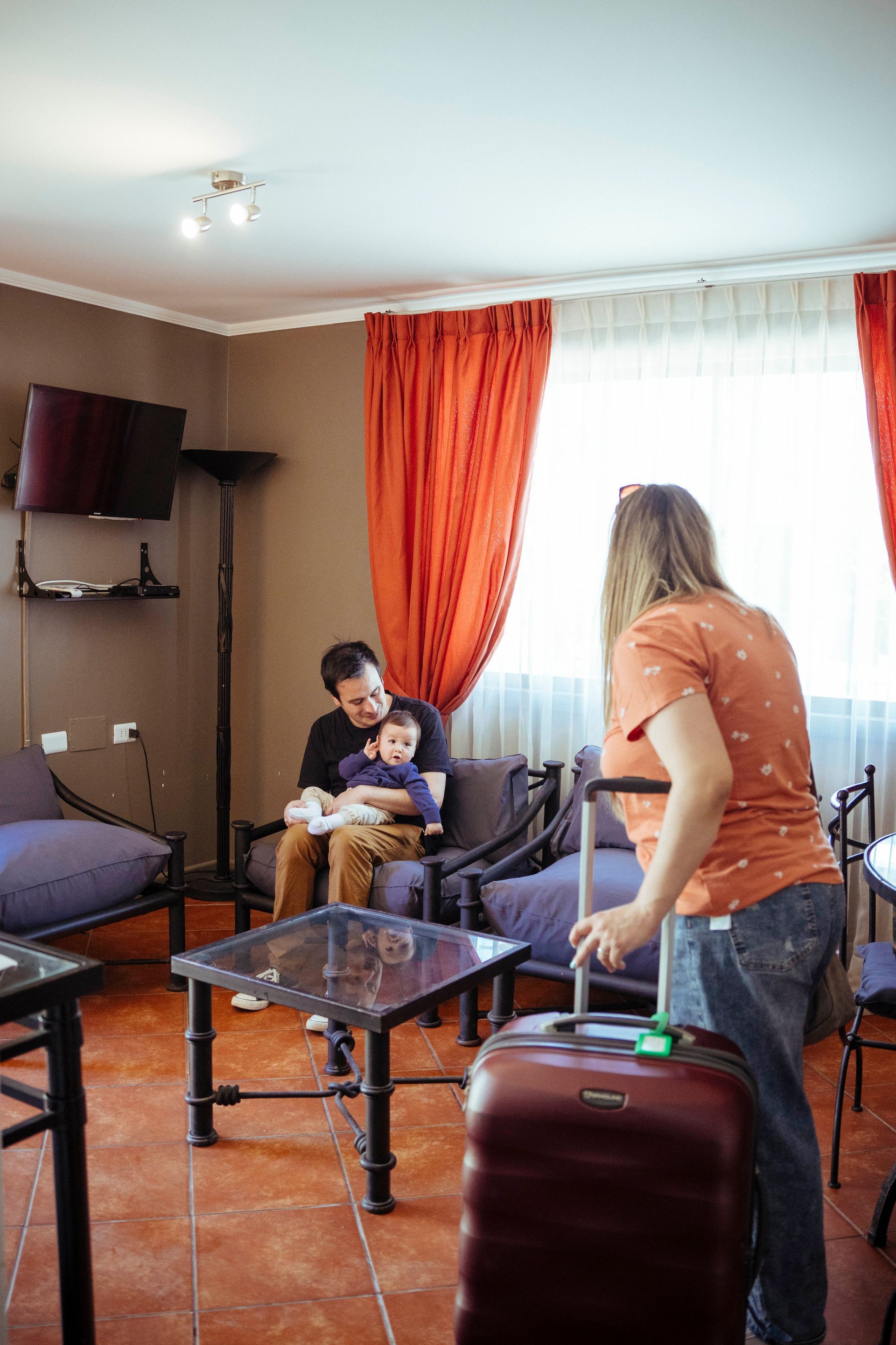 Family in living room, woman with luggage leaving, man holding child. Brown, orange, and blue decor.