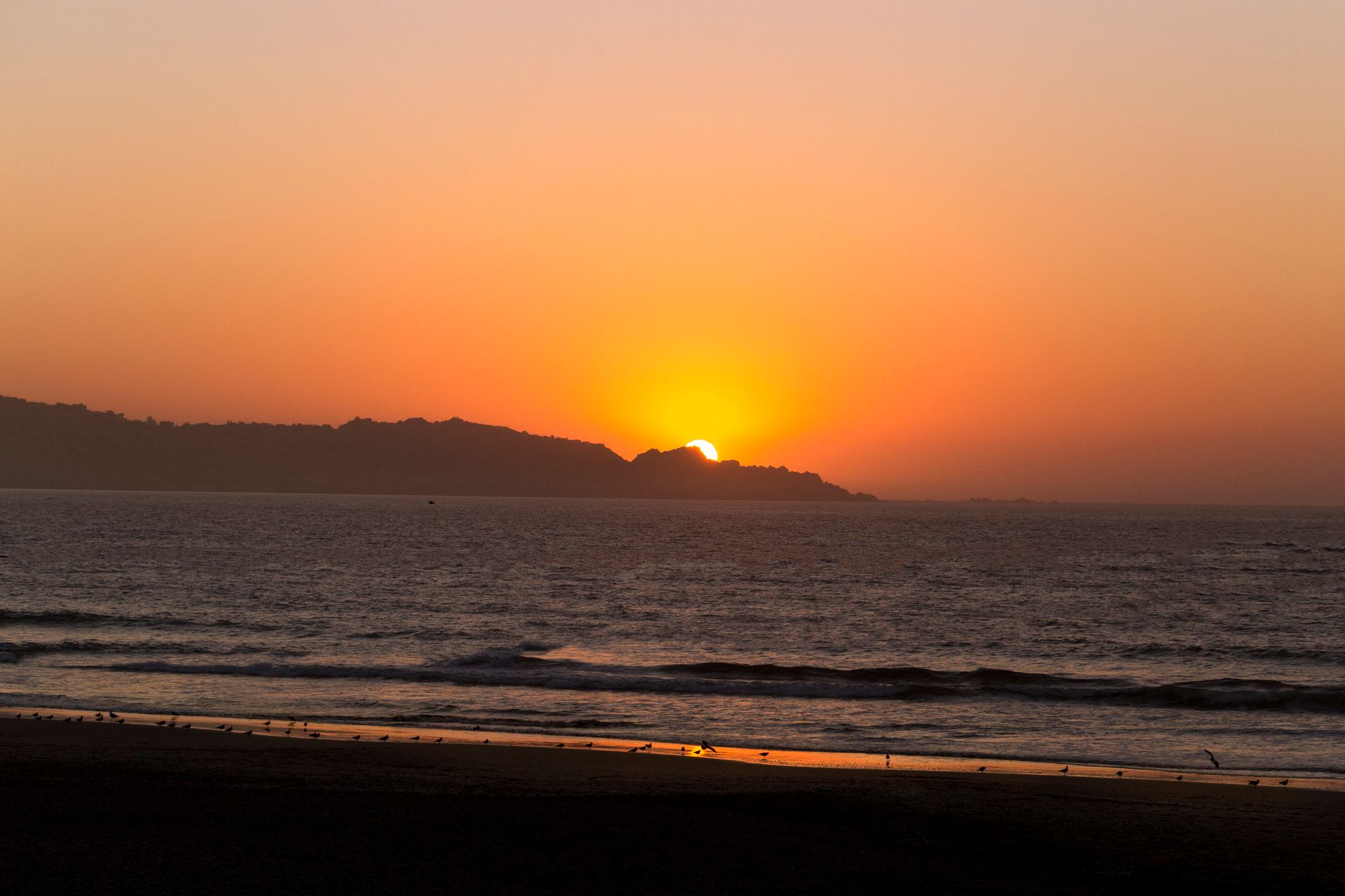Sunset over the ocean with orange sky, distant coastline, and dark waves