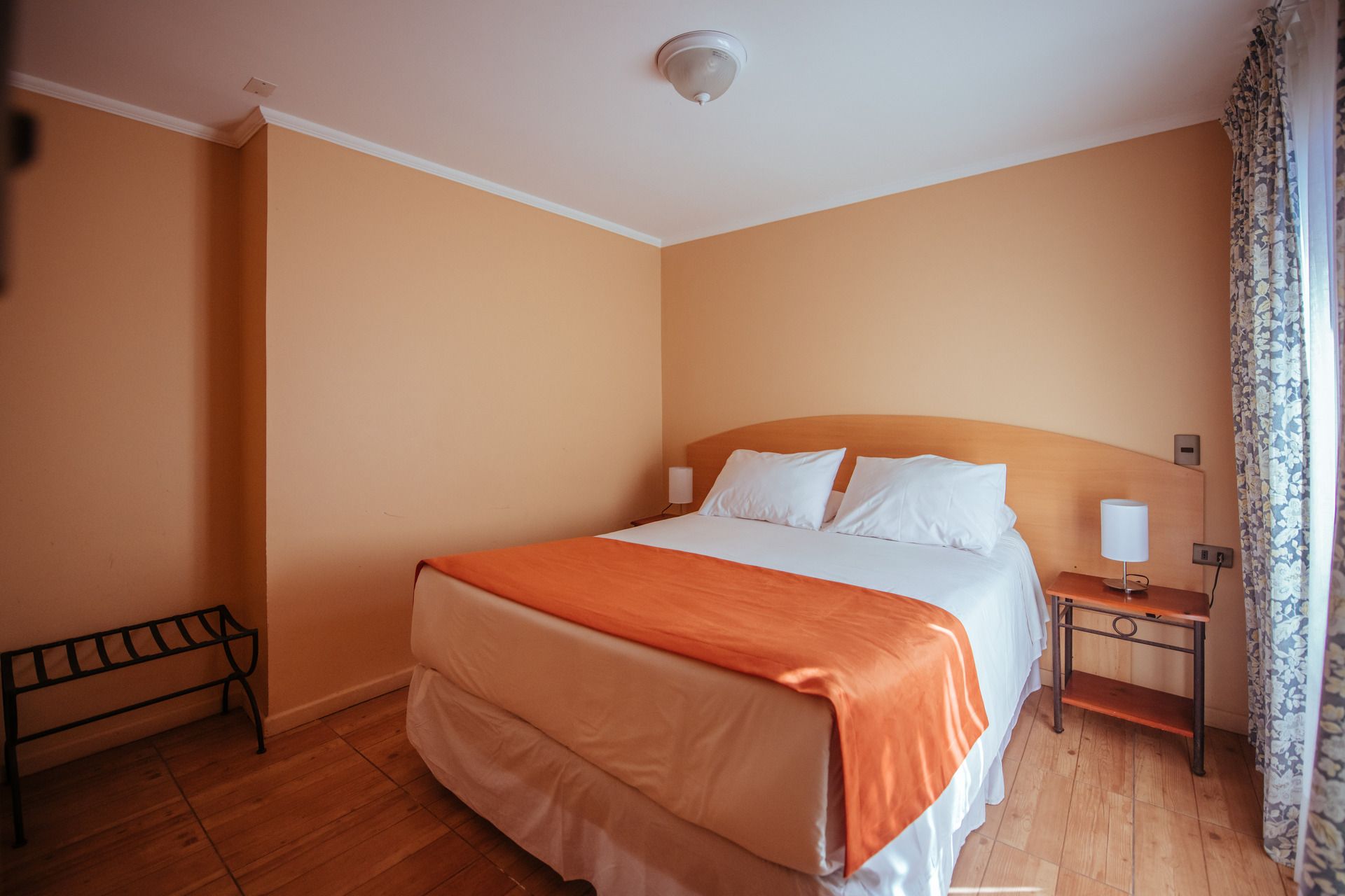 Bedroom with a queen-sized bed, orange blanket, two white pillows, nightstand, luggage rack, and beige walls.