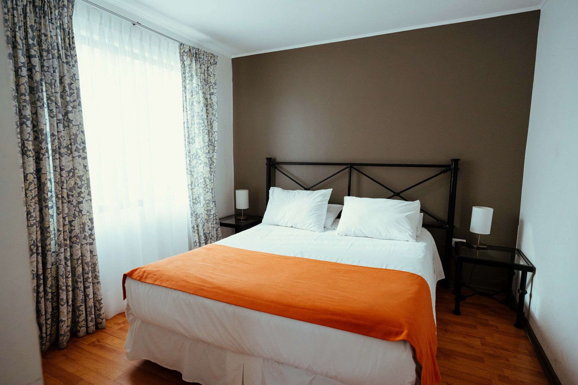 Bright bedroom with a metal bed frame, orange bedspread, white pillows, and large window curtains