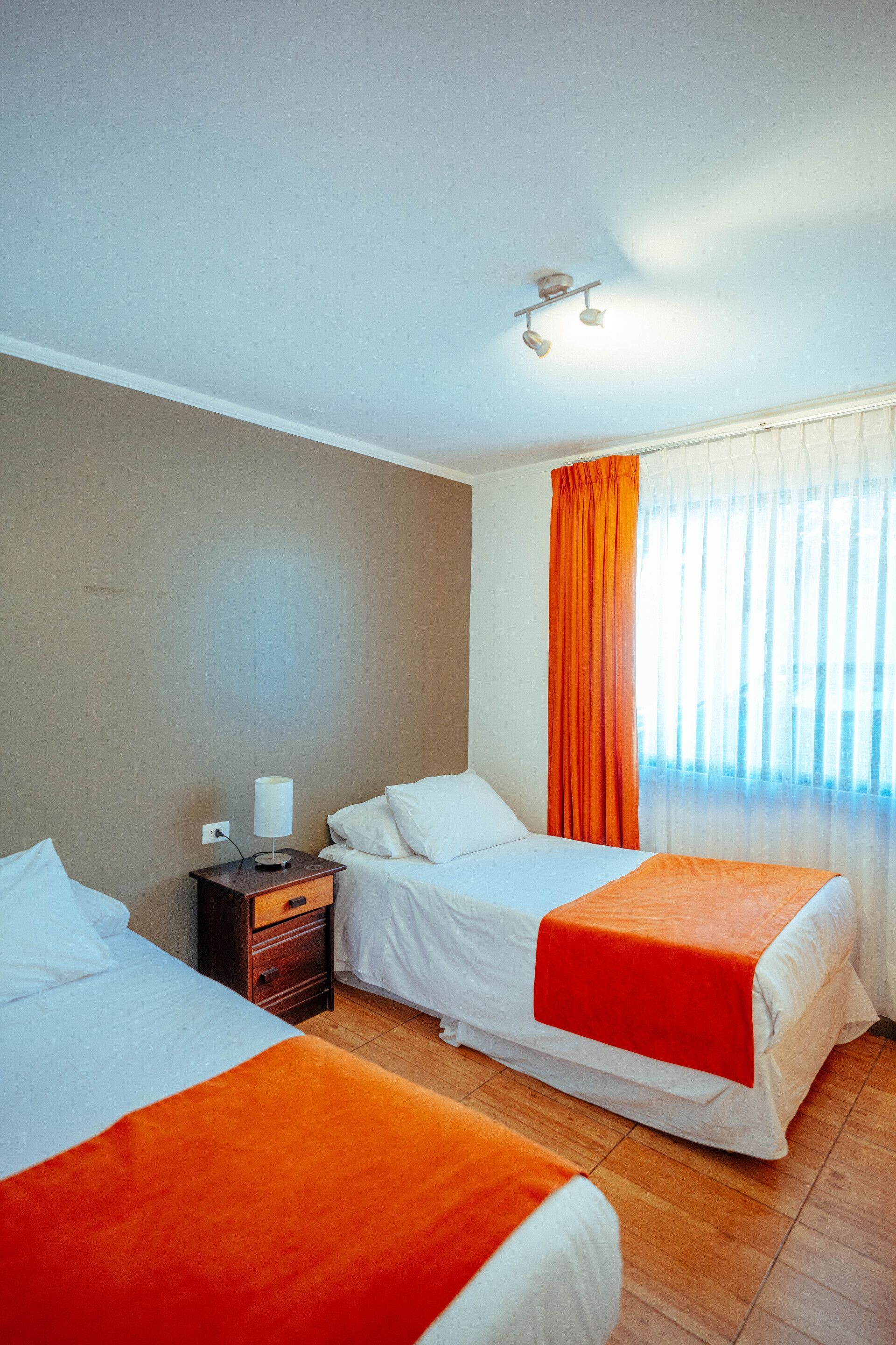 Two beds with orange blankets in a bedroom. Beige and white walls, orange curtain.