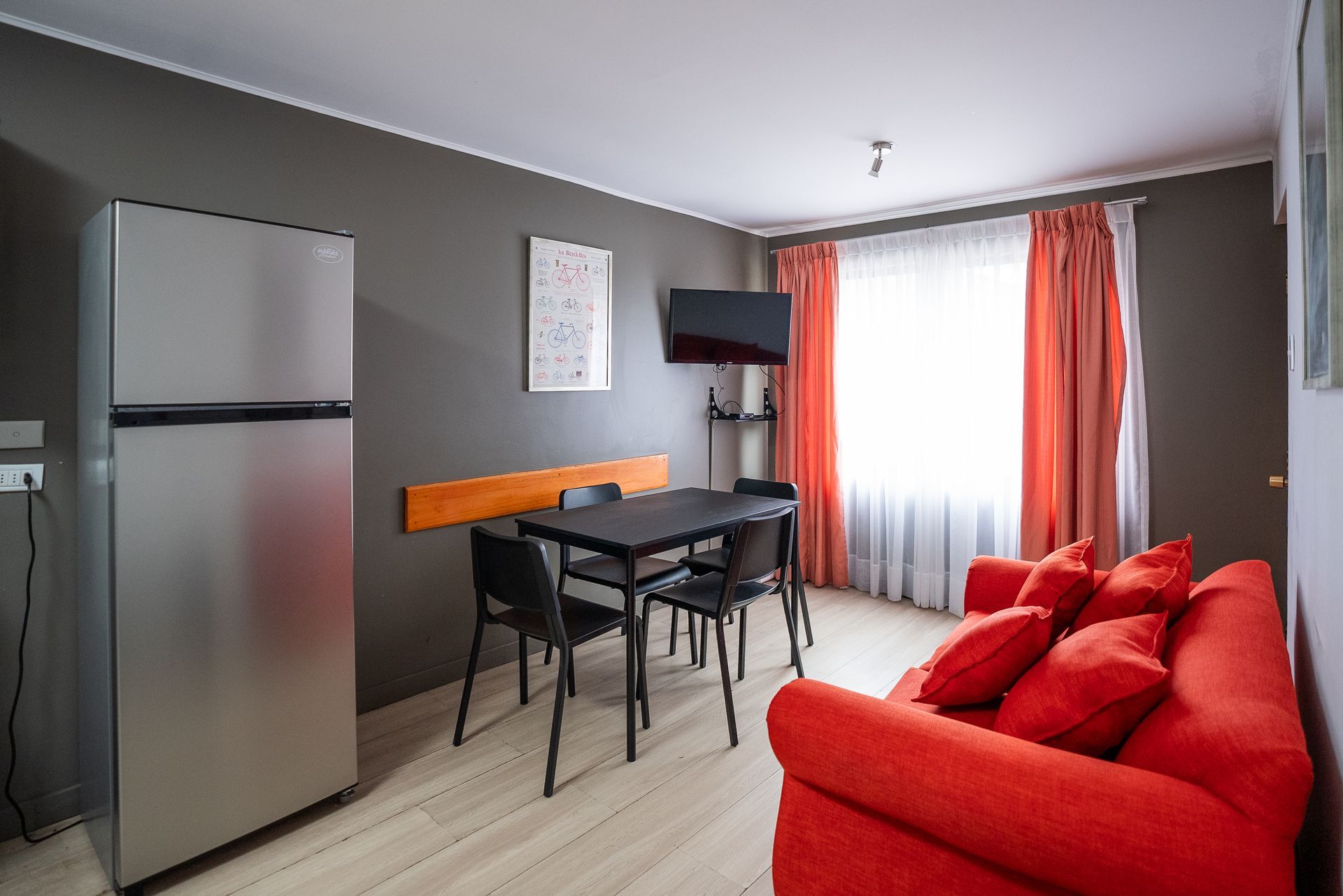 Small modern room with red sofa, dining table, wall-mounted TV, and stainless steel refrigerator.