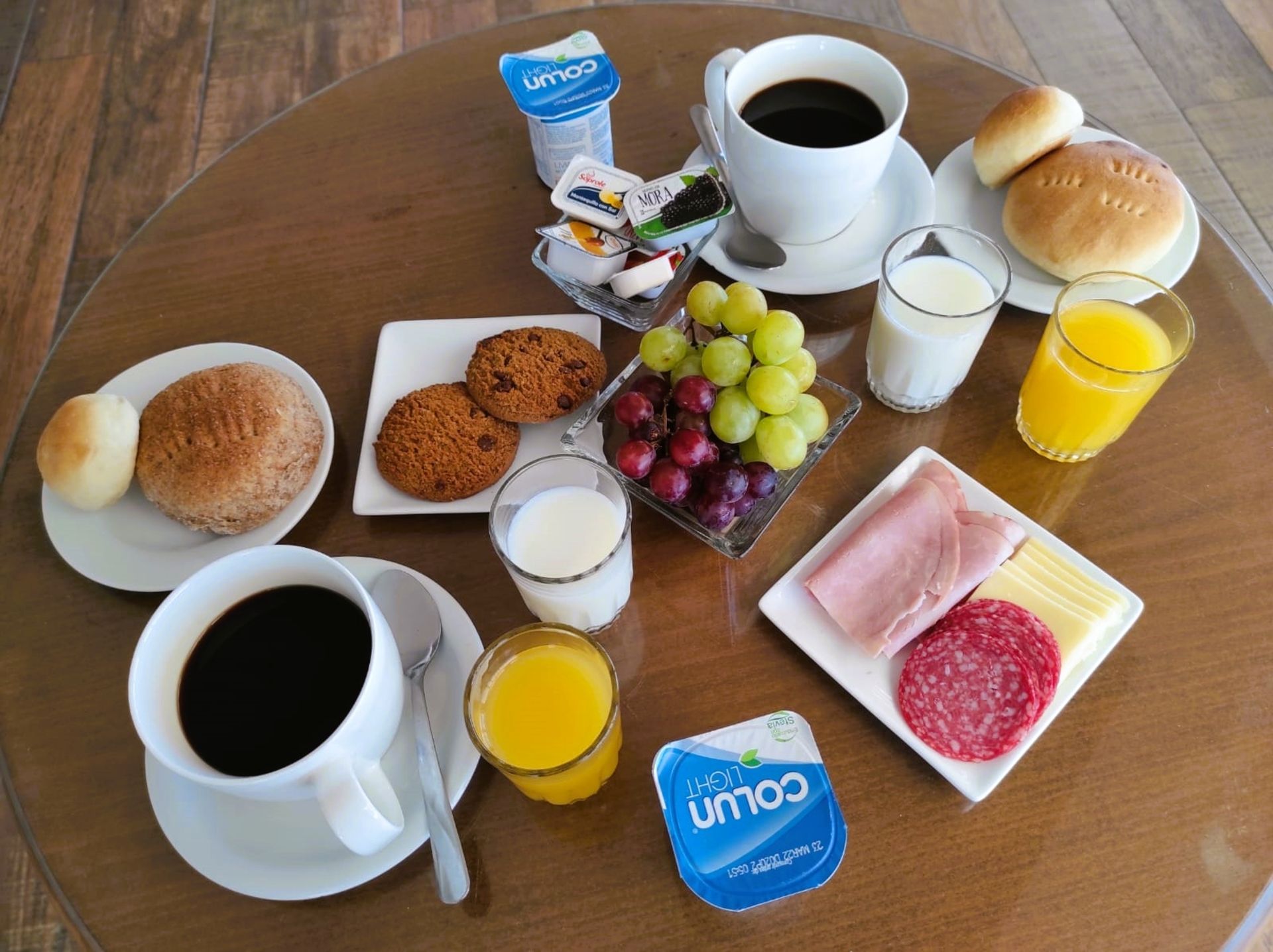 Breakfast table with coffee, orange juice, bread rolls, pastries, grapes, yogurt, and jam.