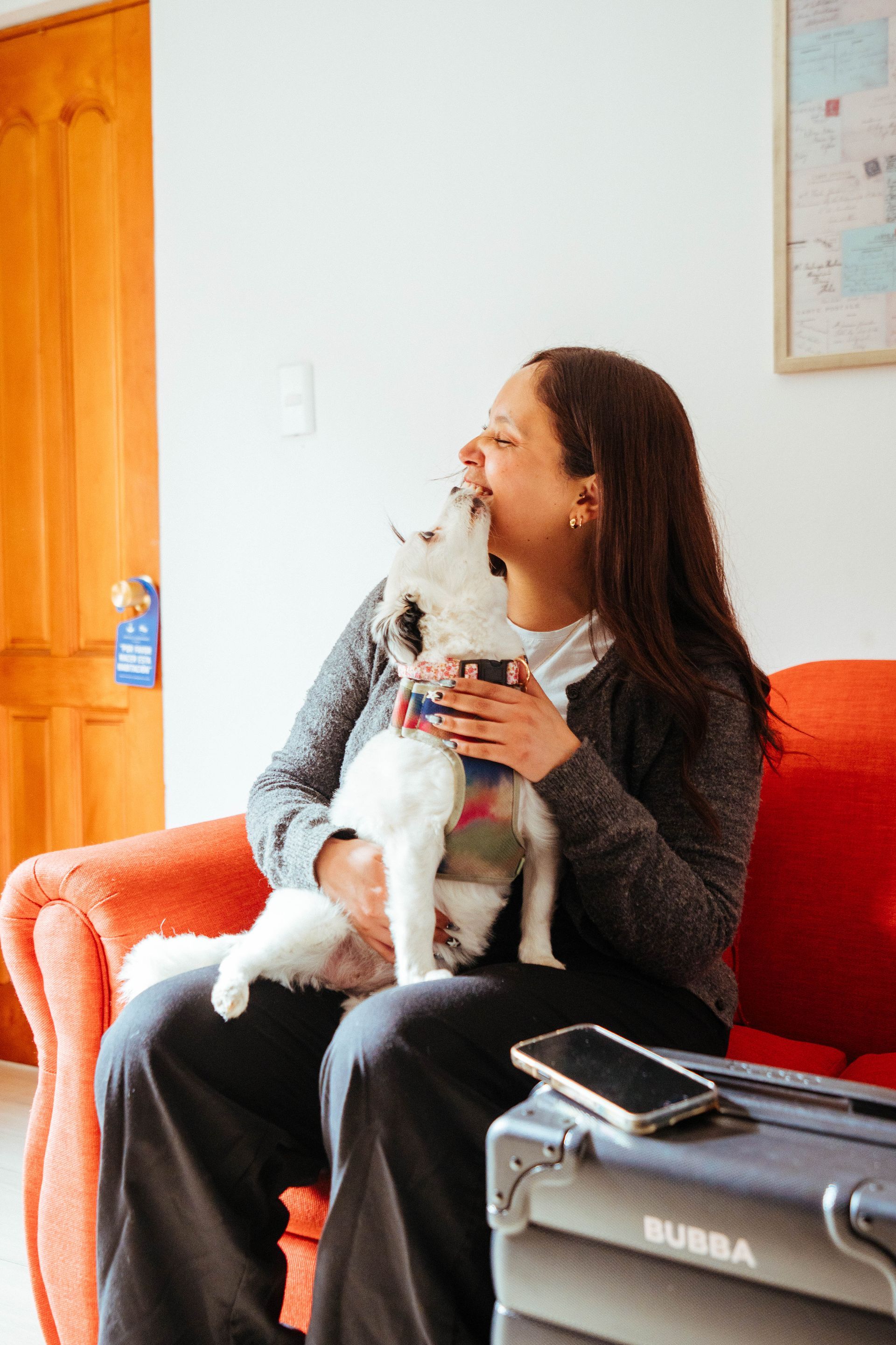 Woman sitting on an orange couch holding a small white dog in a bright room