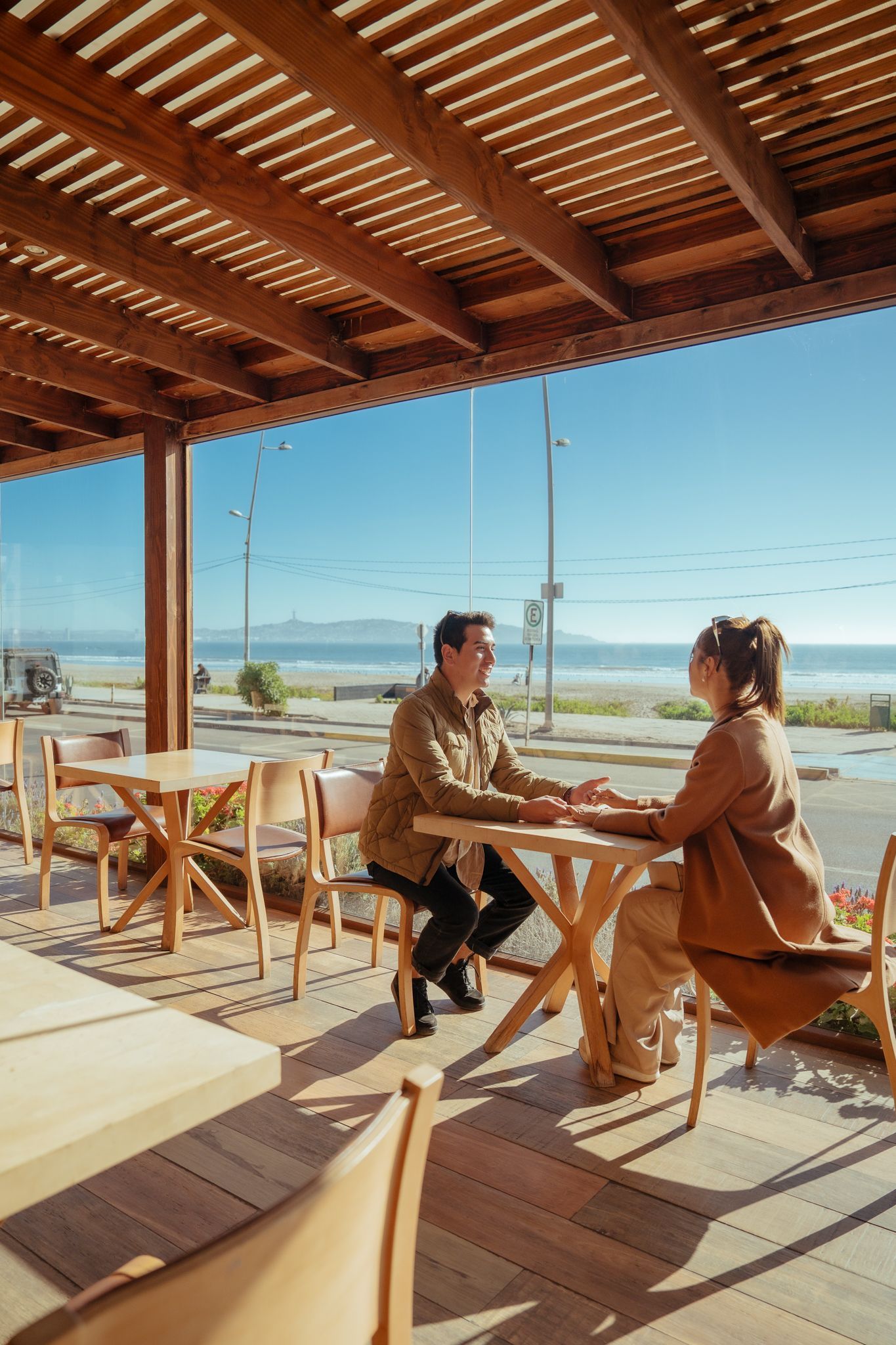 Two people chatting at a seaside café under a wooden pergola, with ocean and blue sky in view