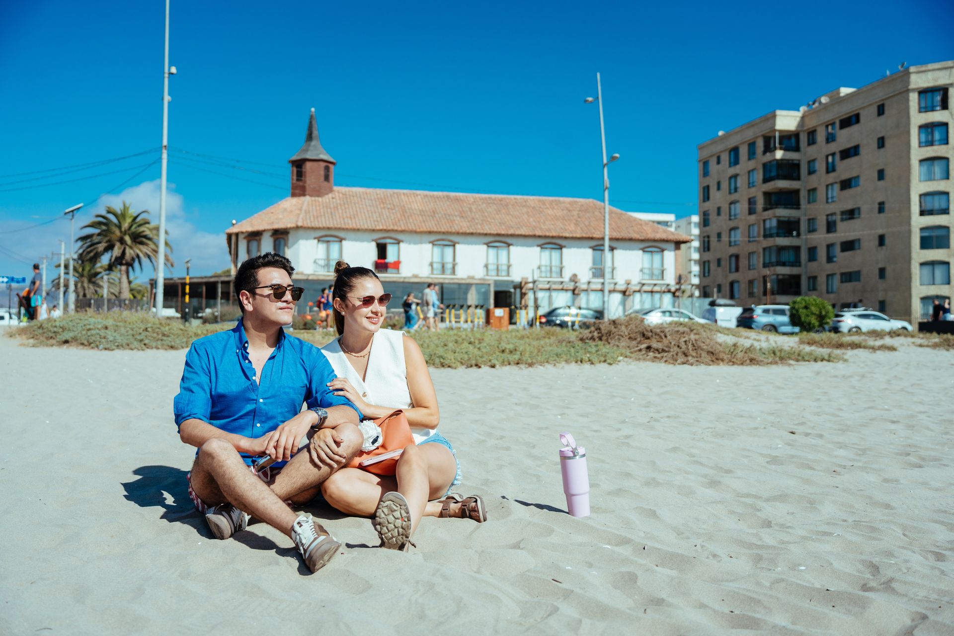 Couple sitting on a sandy beach near a red-roofed building and blue sky