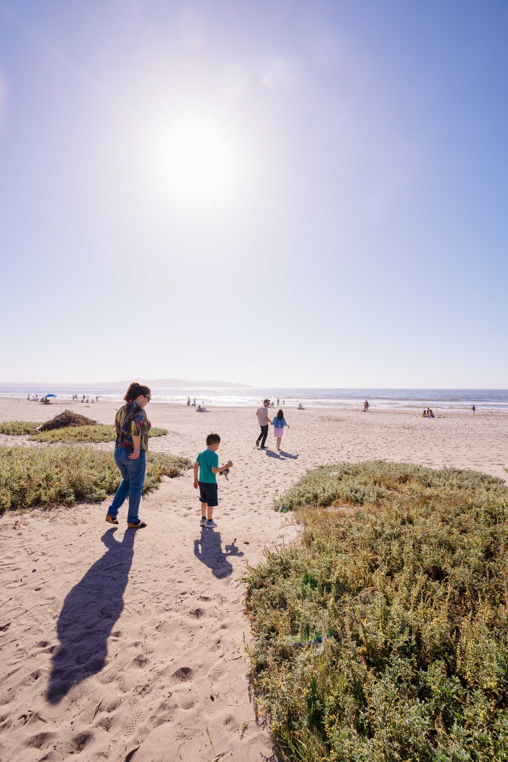 People walking on a sandy beach under bright sun, with low dunes and a wide open sky.