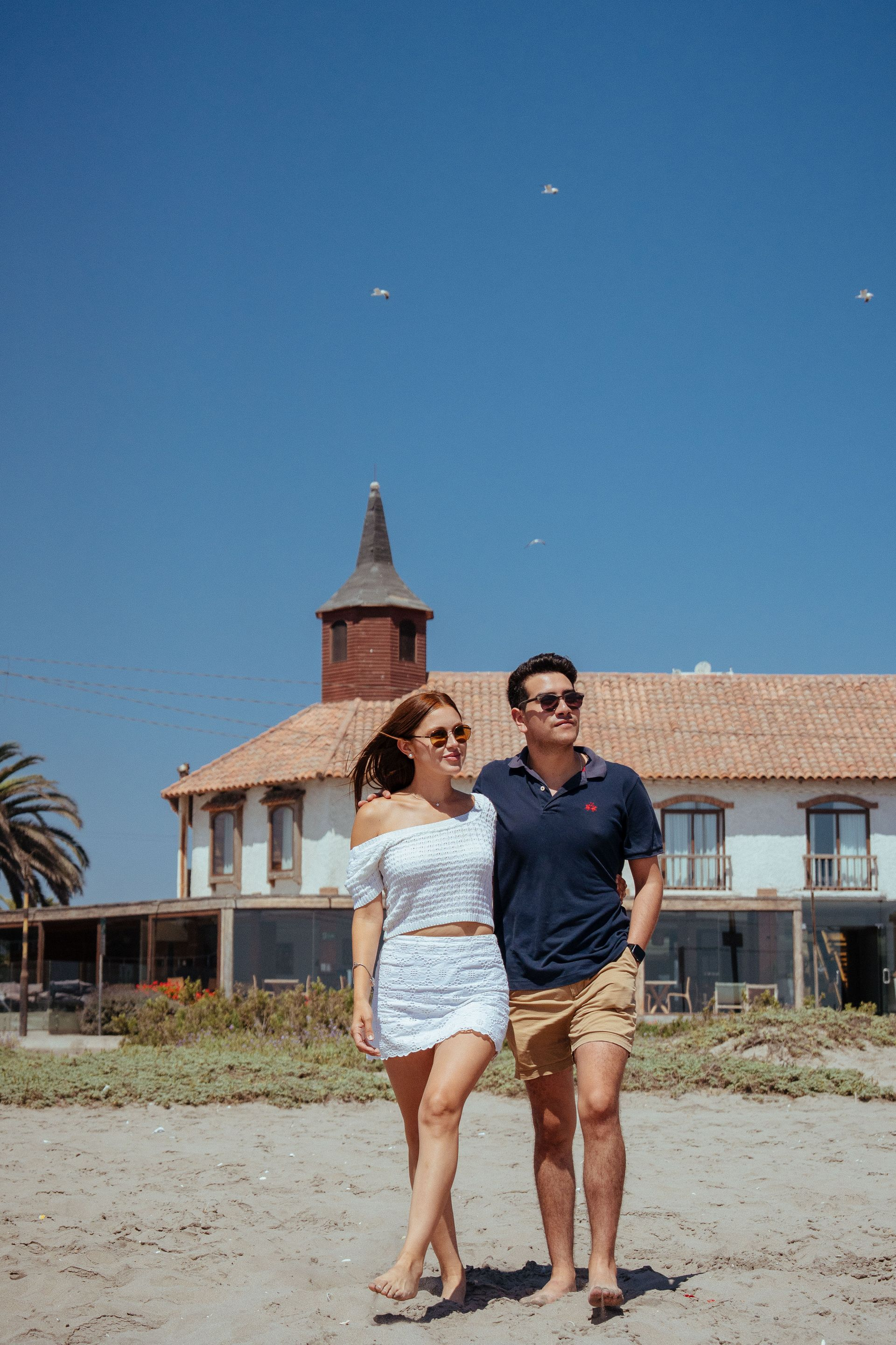 Couple walking on a sandy beach in front of a seaside house under a clear blue sky