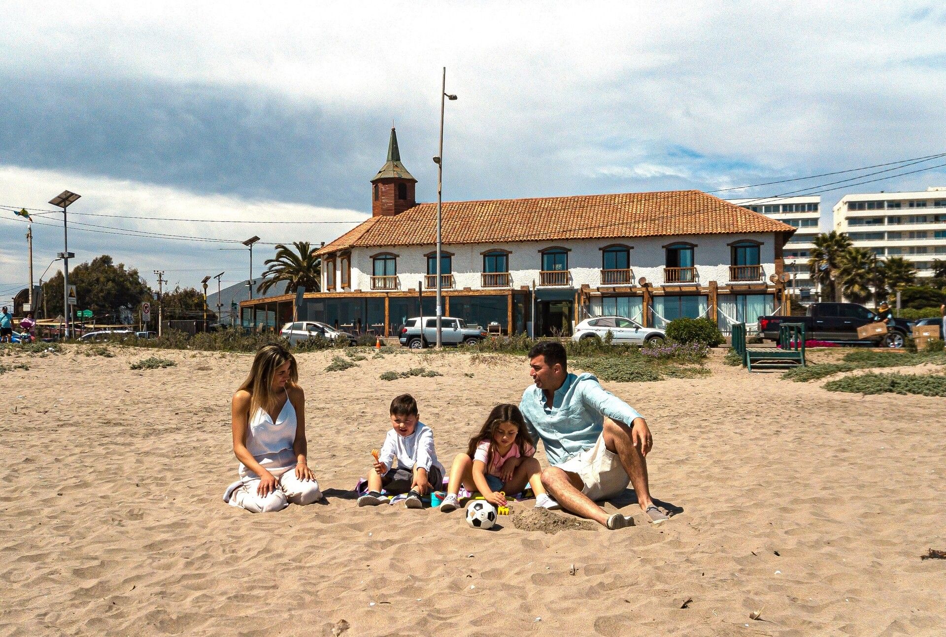 Family sitting on a sandy beach in front of a seaside building under a cloudy sky