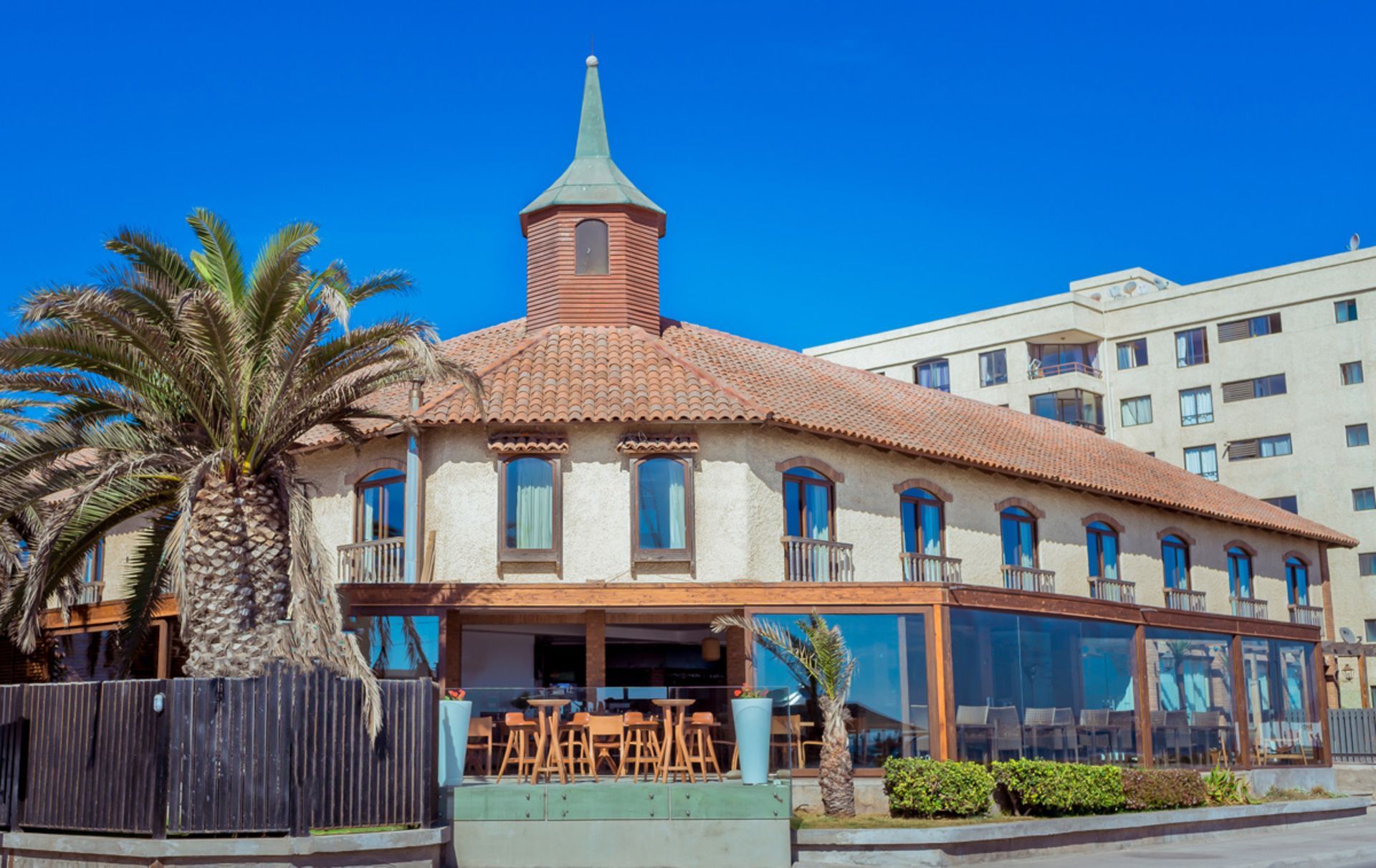 Street-side restaurant with a red-tiled roof and blue awnings beside palm trees under a clear sky