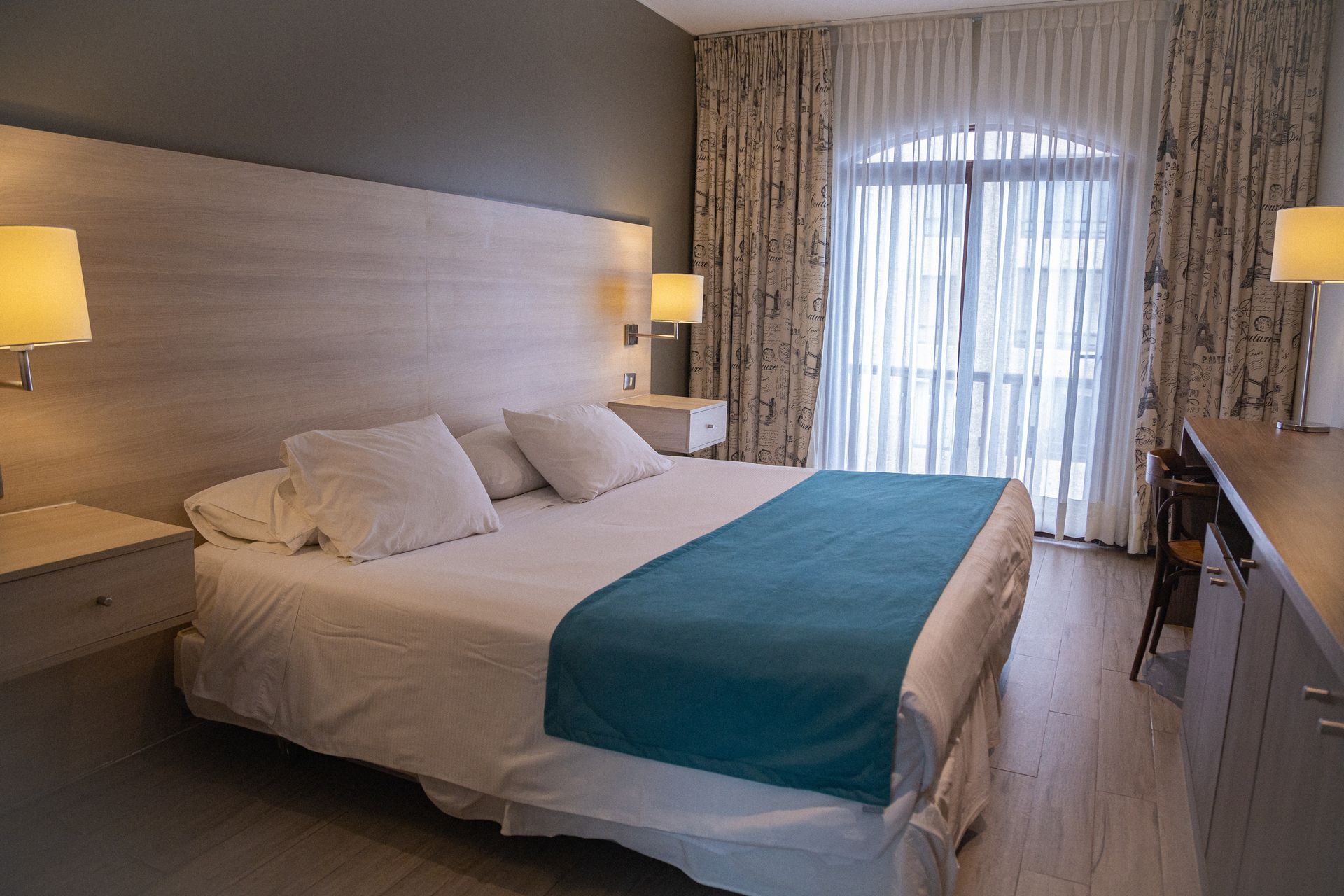 Hotel room with a king-sized bed, nightstands, lamps, and a balcony with sheer curtains.