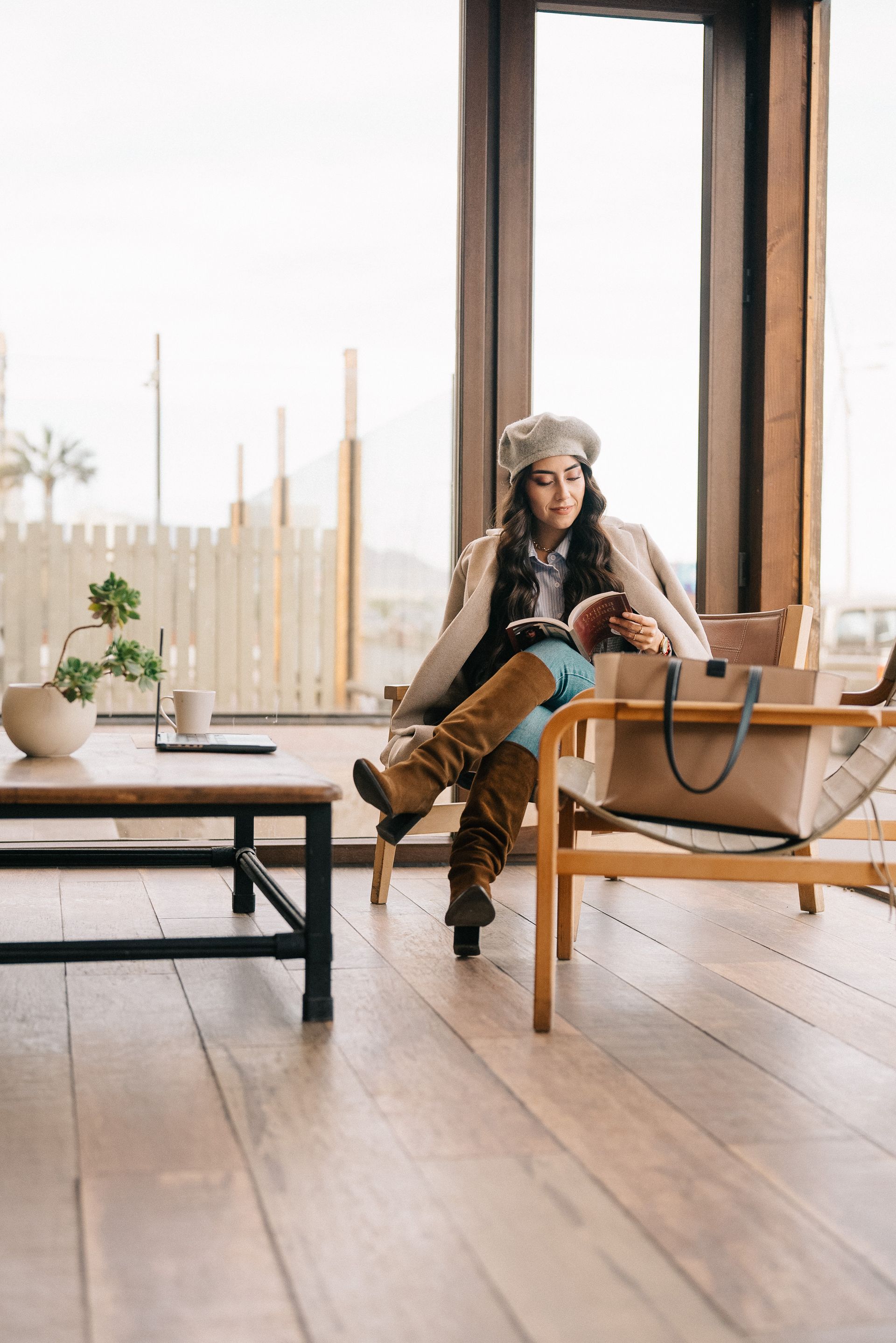 Person sitting on a wooden chair in a bright room with large windows, holding a bag and looking at a phone.