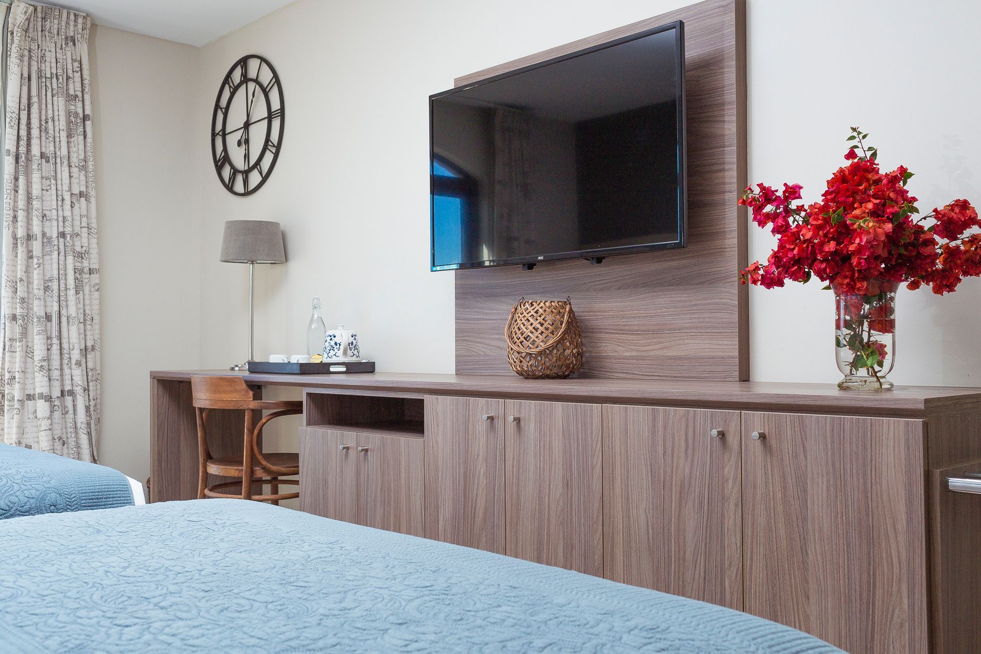 A hotel room with two beds , a desk , a flat screen tv and a vase of flowers.
