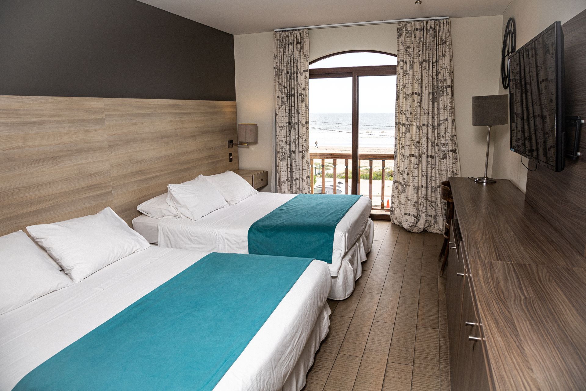 Hotel room with two double beds, teal accents, and a balcony overlooking the ocean.