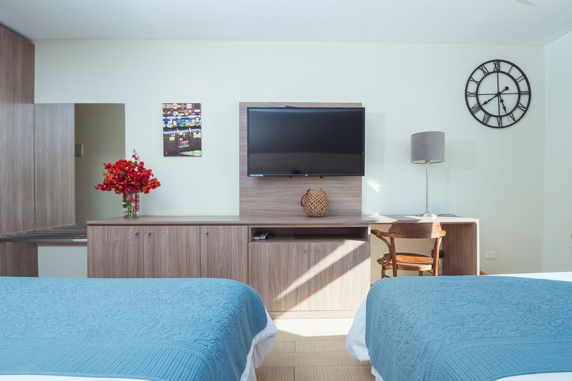 Hotel room with two blue beds, a wall-mounted TV, desk, and a large clock.