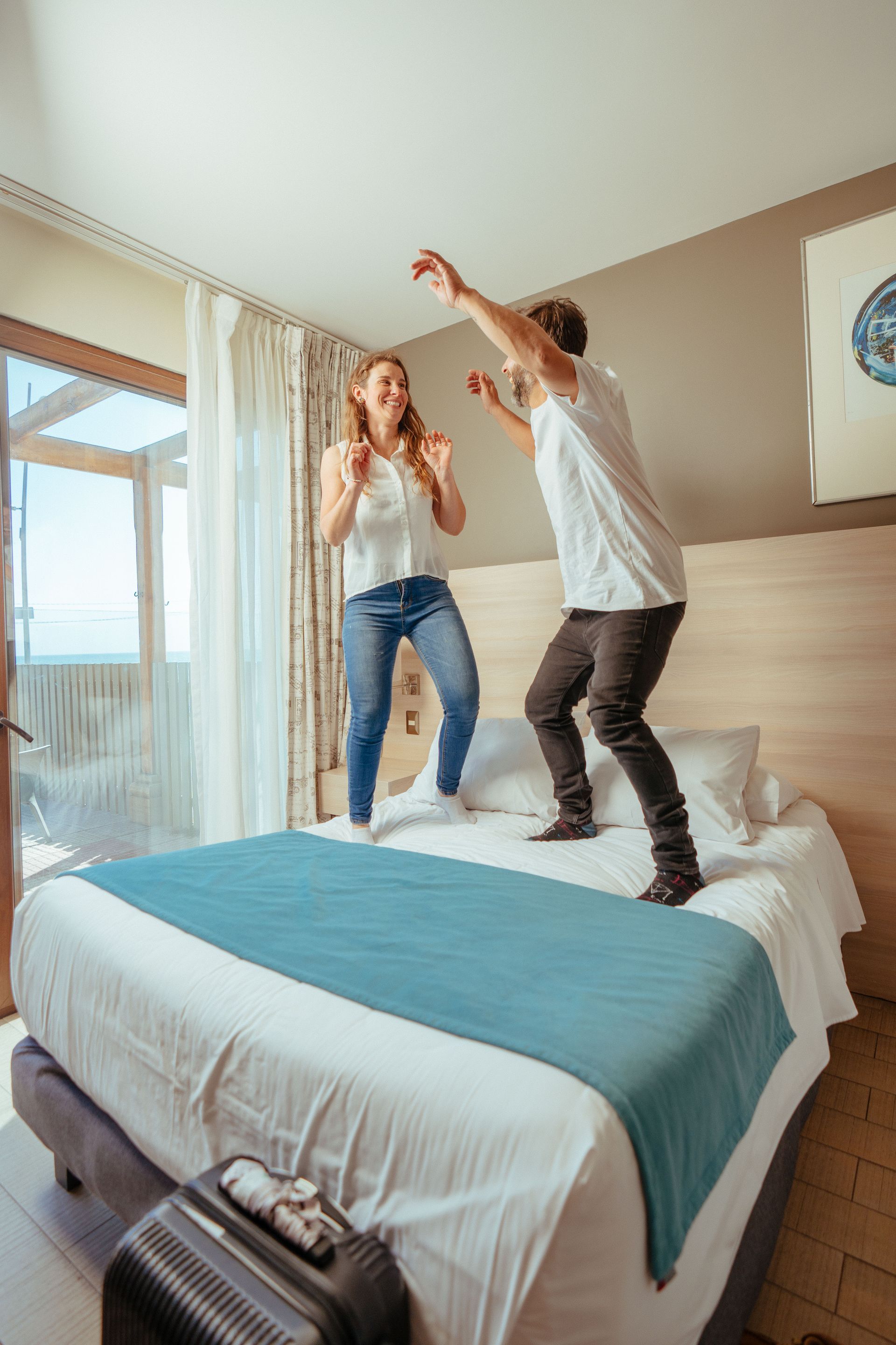 Two people jumping on a bed in a bright hotel room near a window and balcony.