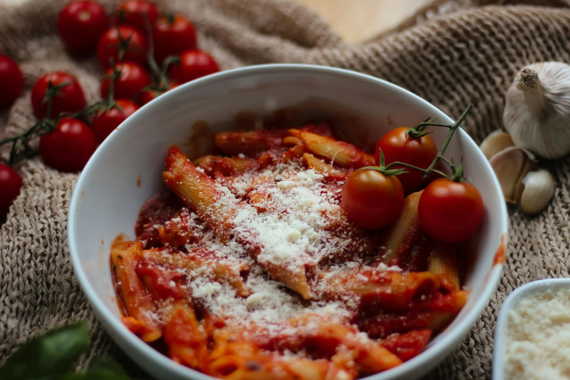 Bowl of pasta with tomato sauce and grated cheese, surrounded by cherry tomatoes and herbs on a table