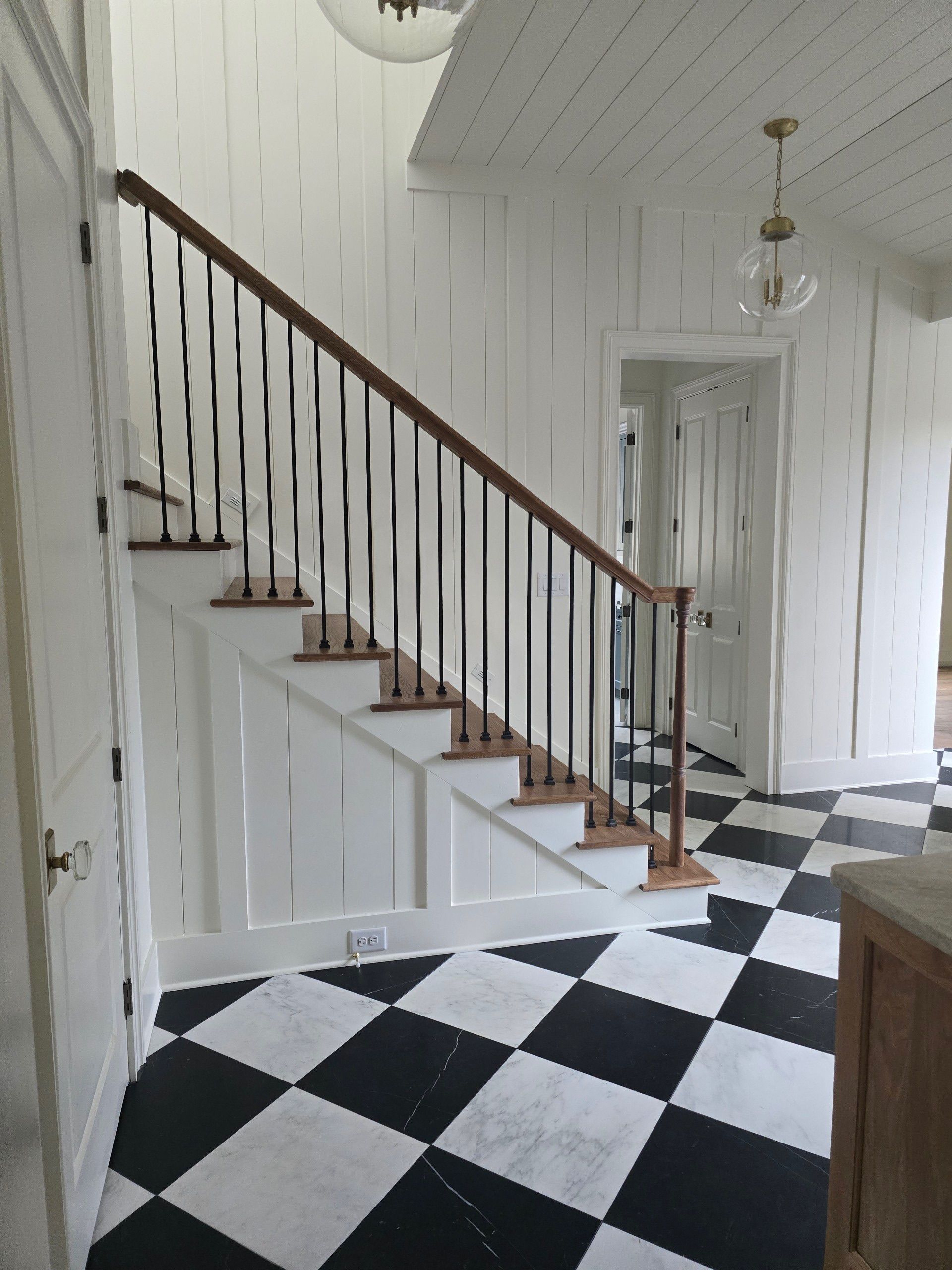 Staircase with wooden steps and black railing in a room with black and white checkered floor and white paneled walls.