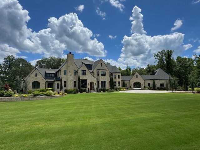 Large stone mansion with a green lawn under a blue sky with fluffy white clouds.