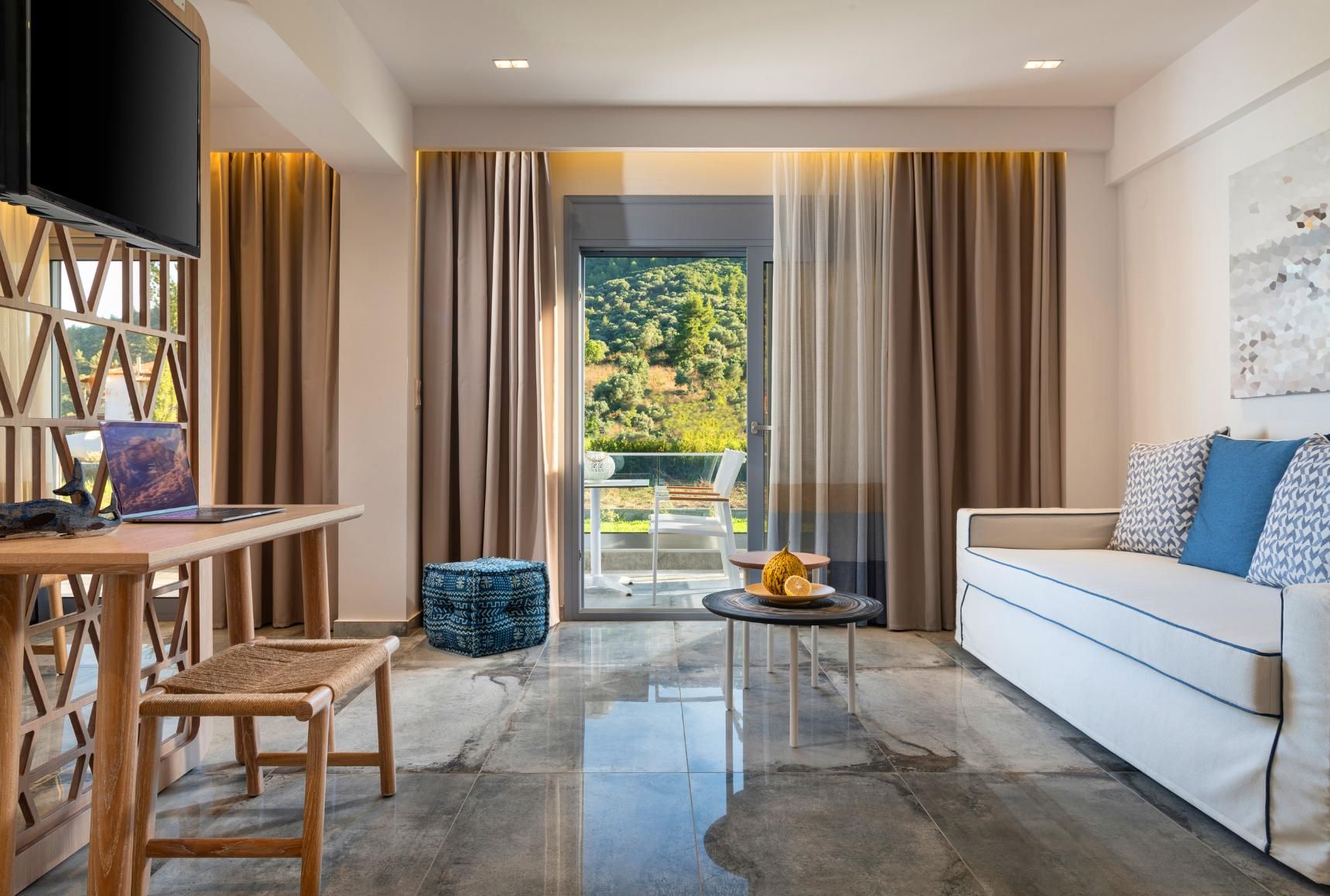 Living room with desk, sofa, and balcony access. Earth-toned curtains, gray flooring, and a blue patterned ottoman.