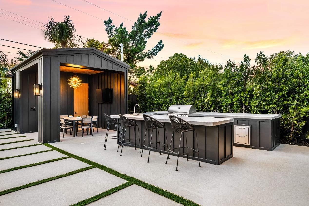 Outdoor kitchen with a grill, bar seating, and dining area under a dark-colored shed; lush greenery in background.