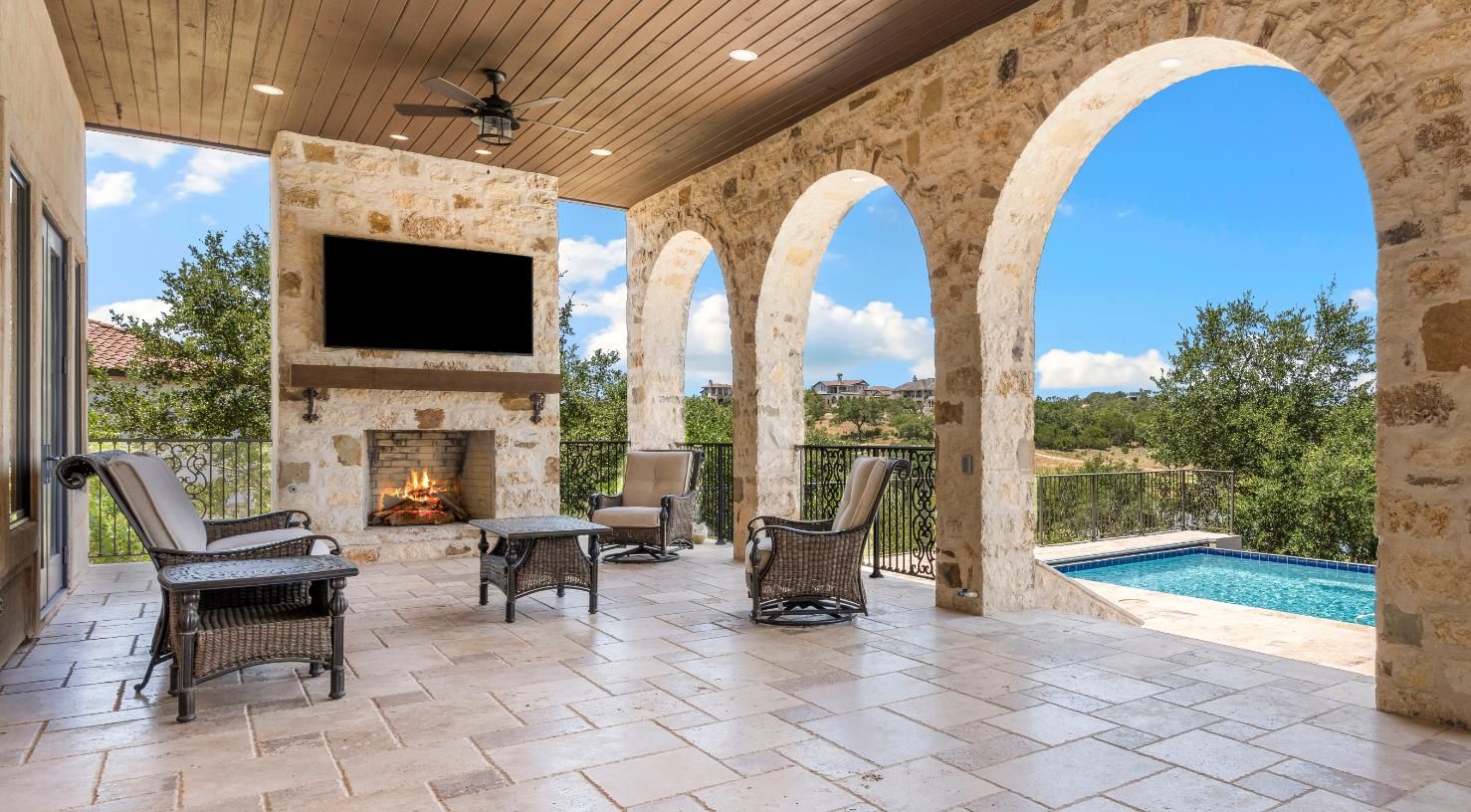 Outdoor patio with stone arches, fireplace, television, seating, and a pool view.