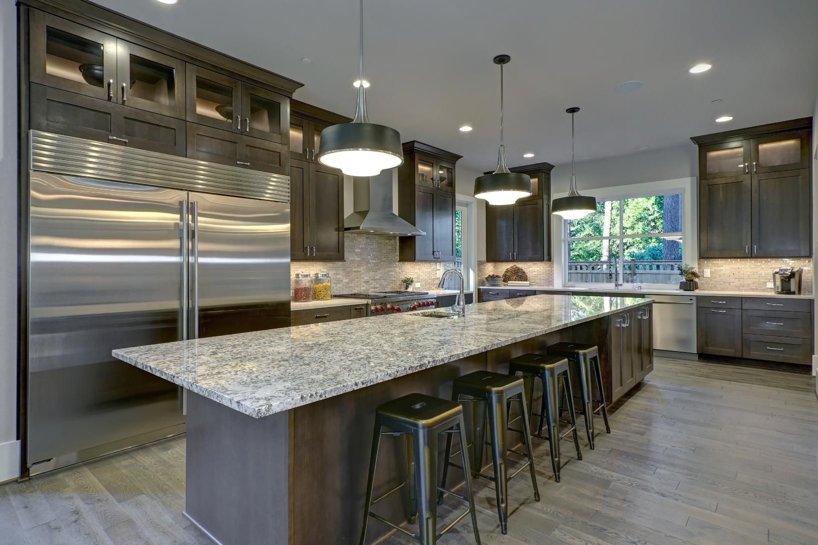 Modern kitchen with large island, stainless steel refrigerator, dark wood cabinets, granite countertops, and pendant lights.