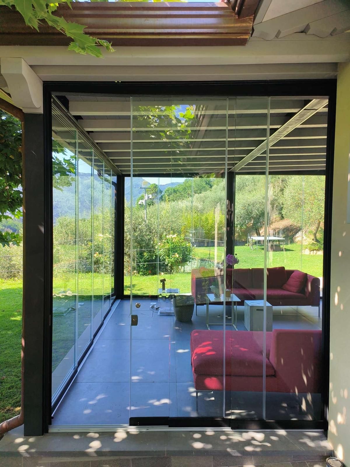 Patio con vetrate e divani rossi, con vista su un giardino verde con alberi e montagne.