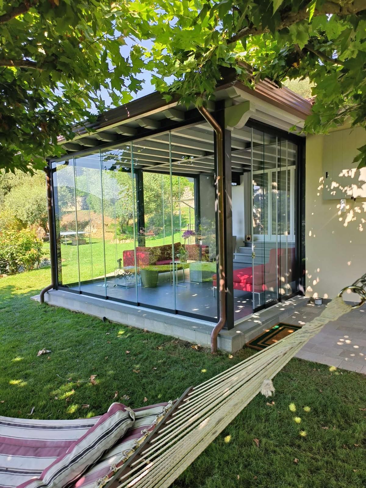 Patio con vetrate e amaca di fronte; sedute rosse visibili all'interno, circondato da prato verde e alberi.
