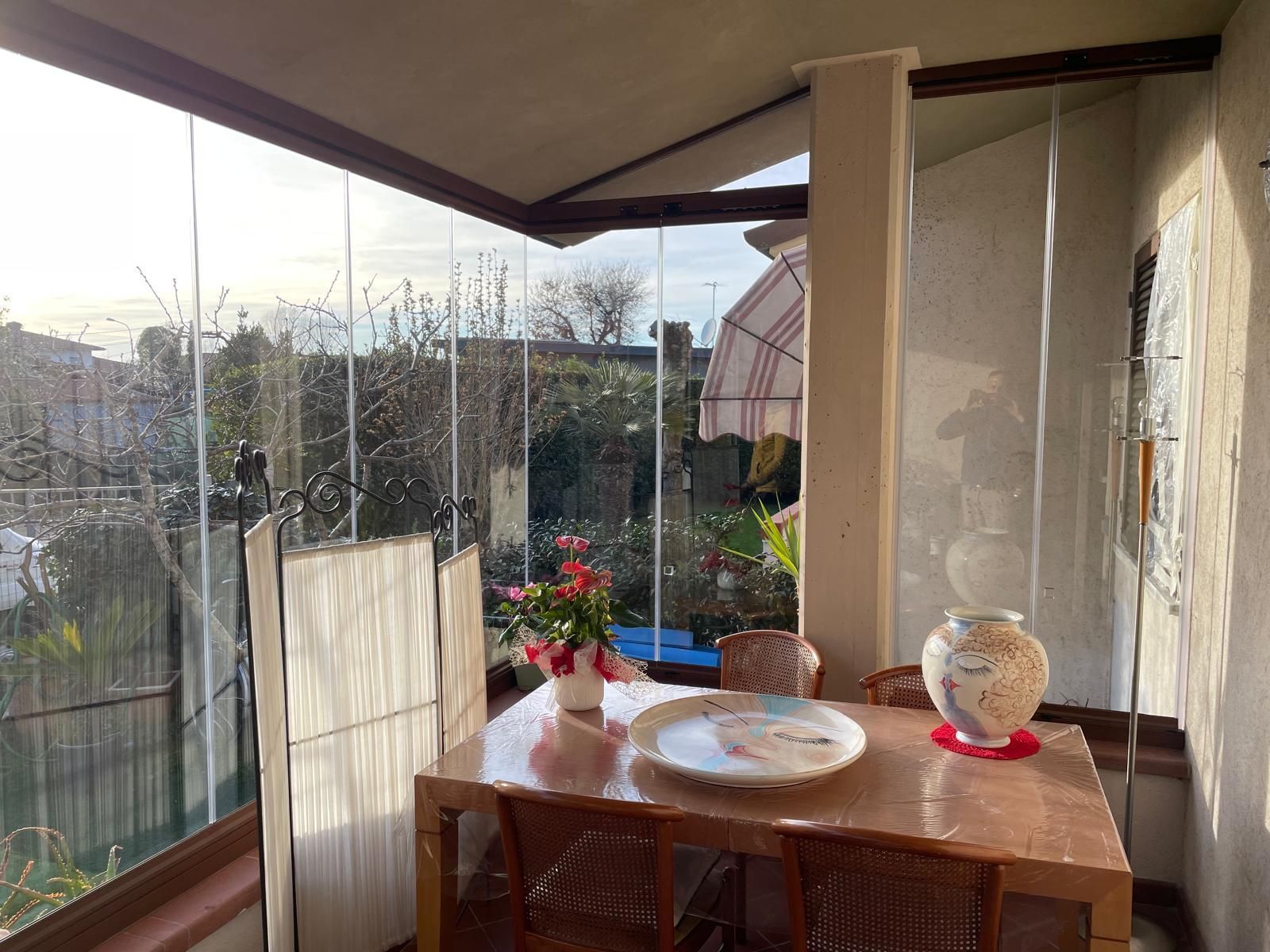 Patio soleggiato e recintato con pareti in vetro, tavolo apparecchiato con vaso e fiori, con vista sul giardino.