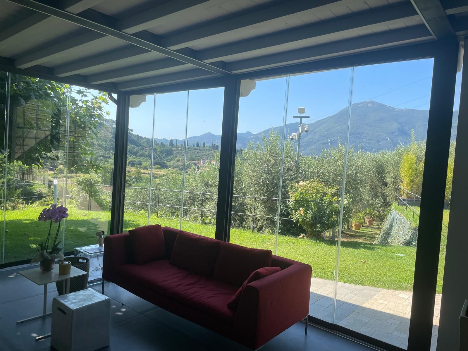 Divano rosso in una veranda con pareti in vetro che si affaccia sulle montagne e sul verde in una giornata di sole.